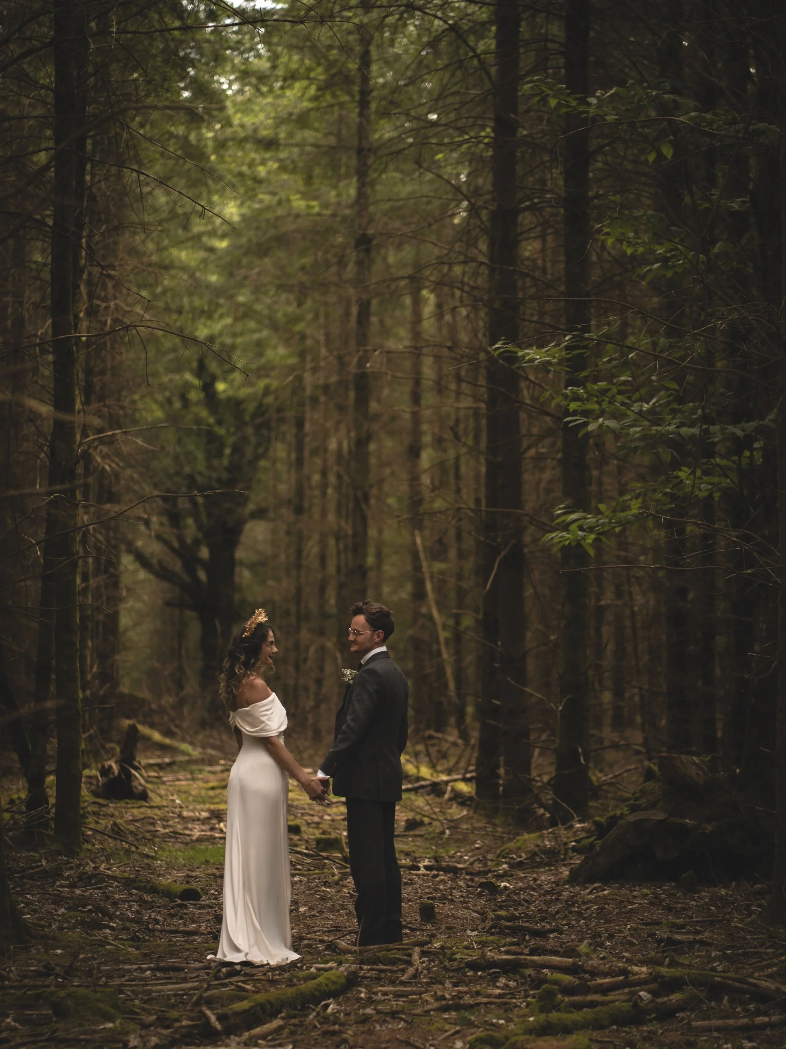 A couple dressed in wedding attire holding hands and standing in a forested area, facing each other and smiling.
