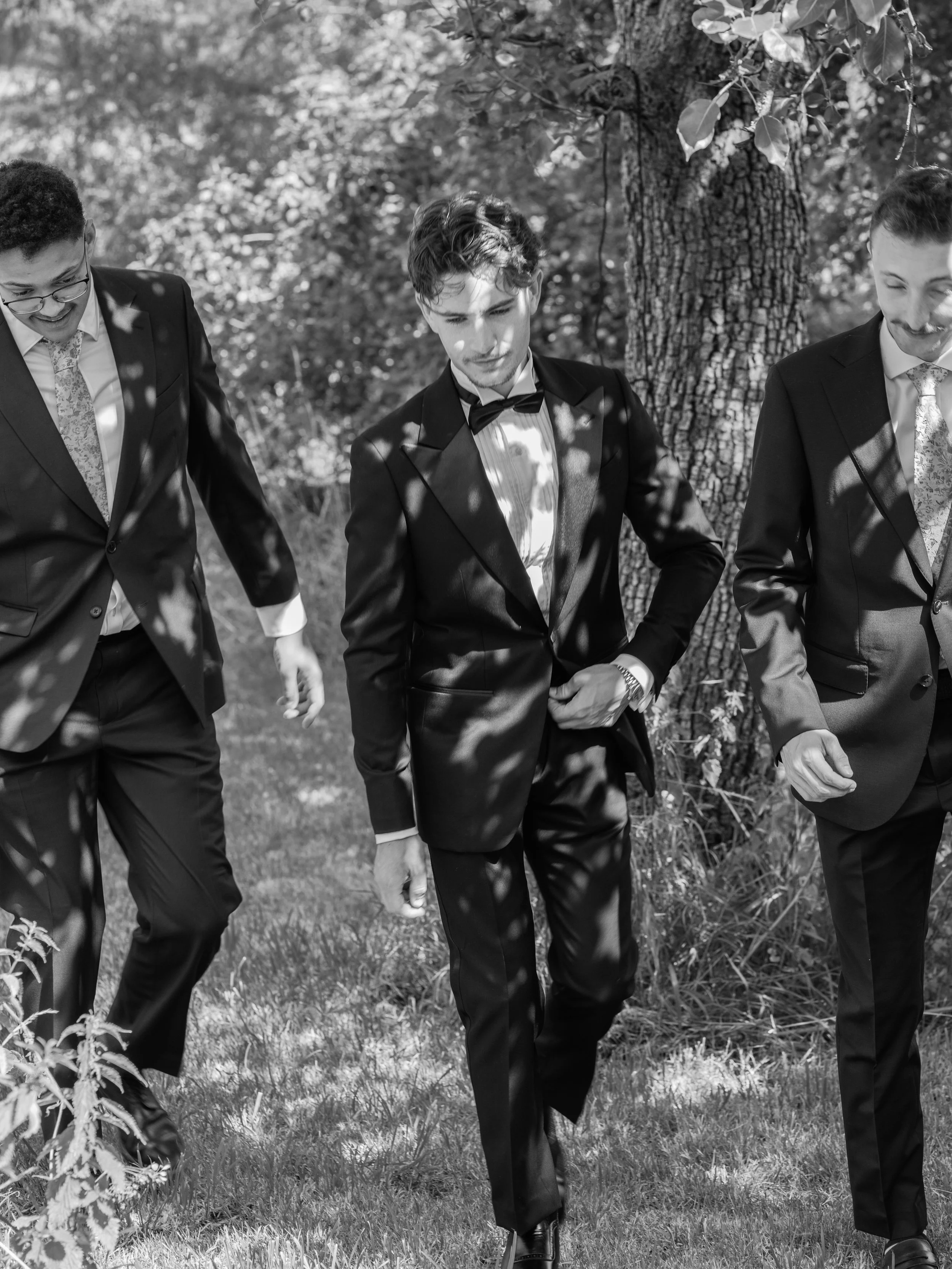Three men dressed in tuxedos walking outdoors on grass, with trees in the background.
