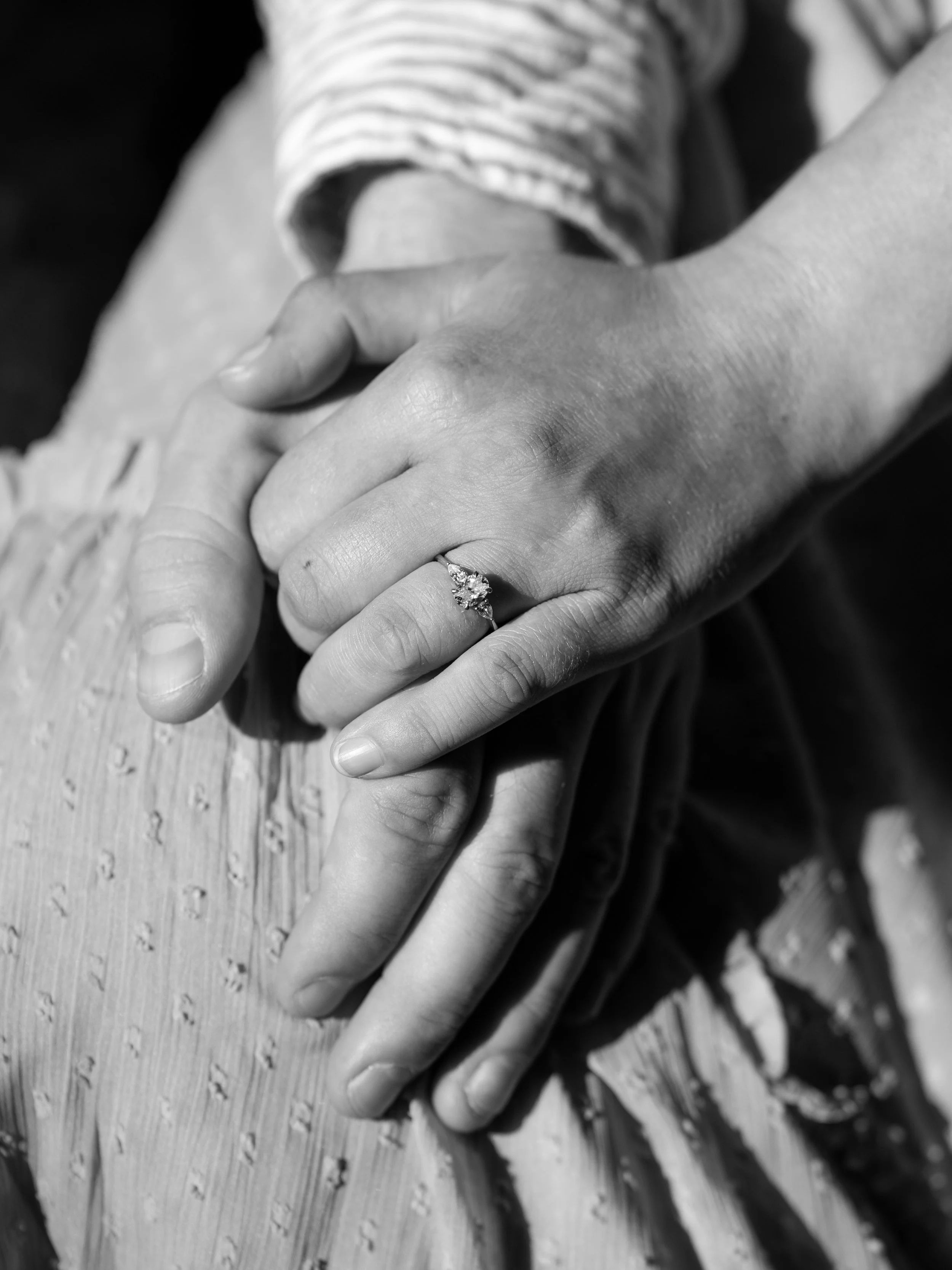 Close-up of two hands gently holding each other, one of which has an engagement ring with a central diamond and smaller side stones, on a wooden surface background.