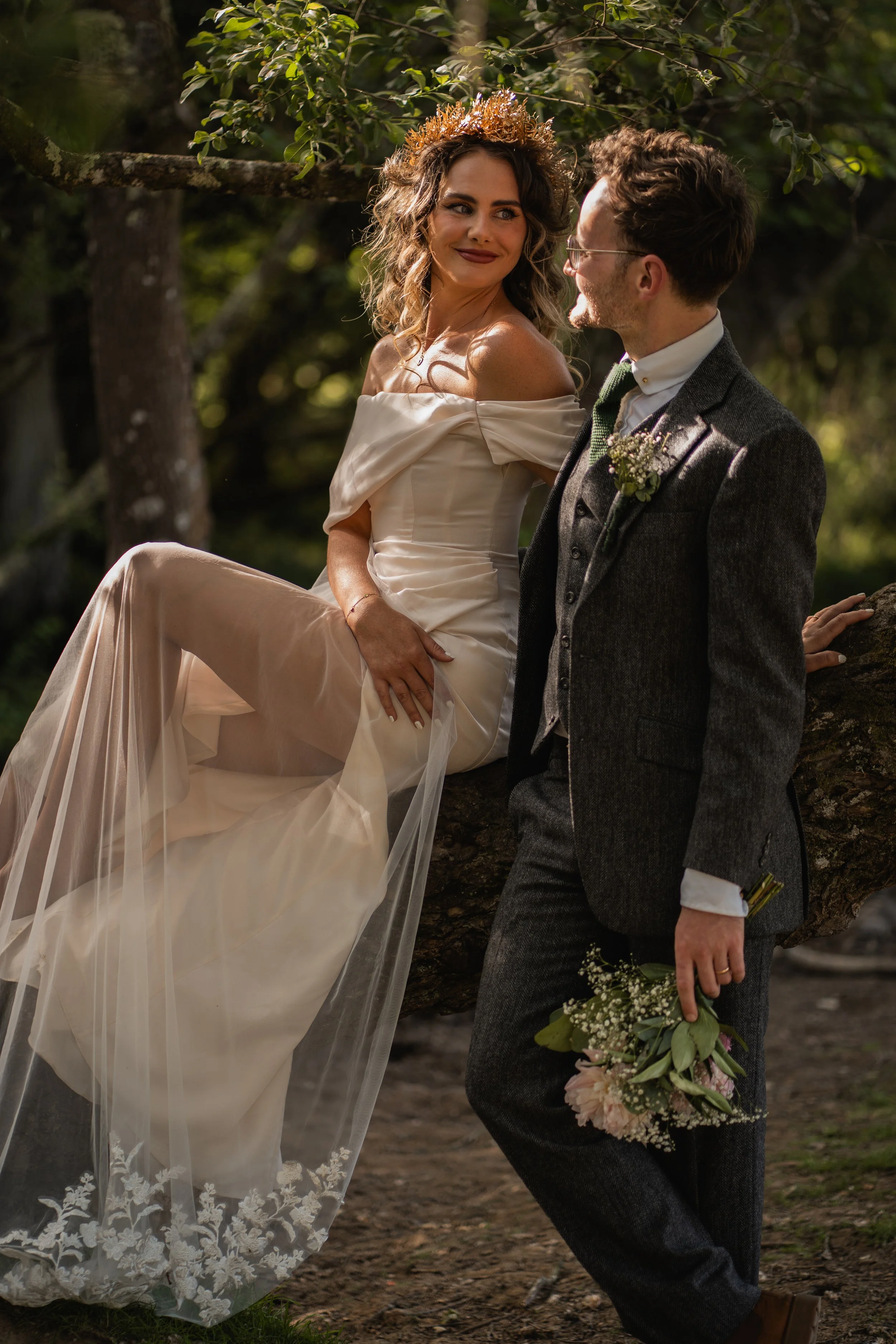 A bride and groom in wedding attire outdoors, with the bride sitting on a tree branch and the groom standing beside her, holding a bouquet, surrounded by greenery.