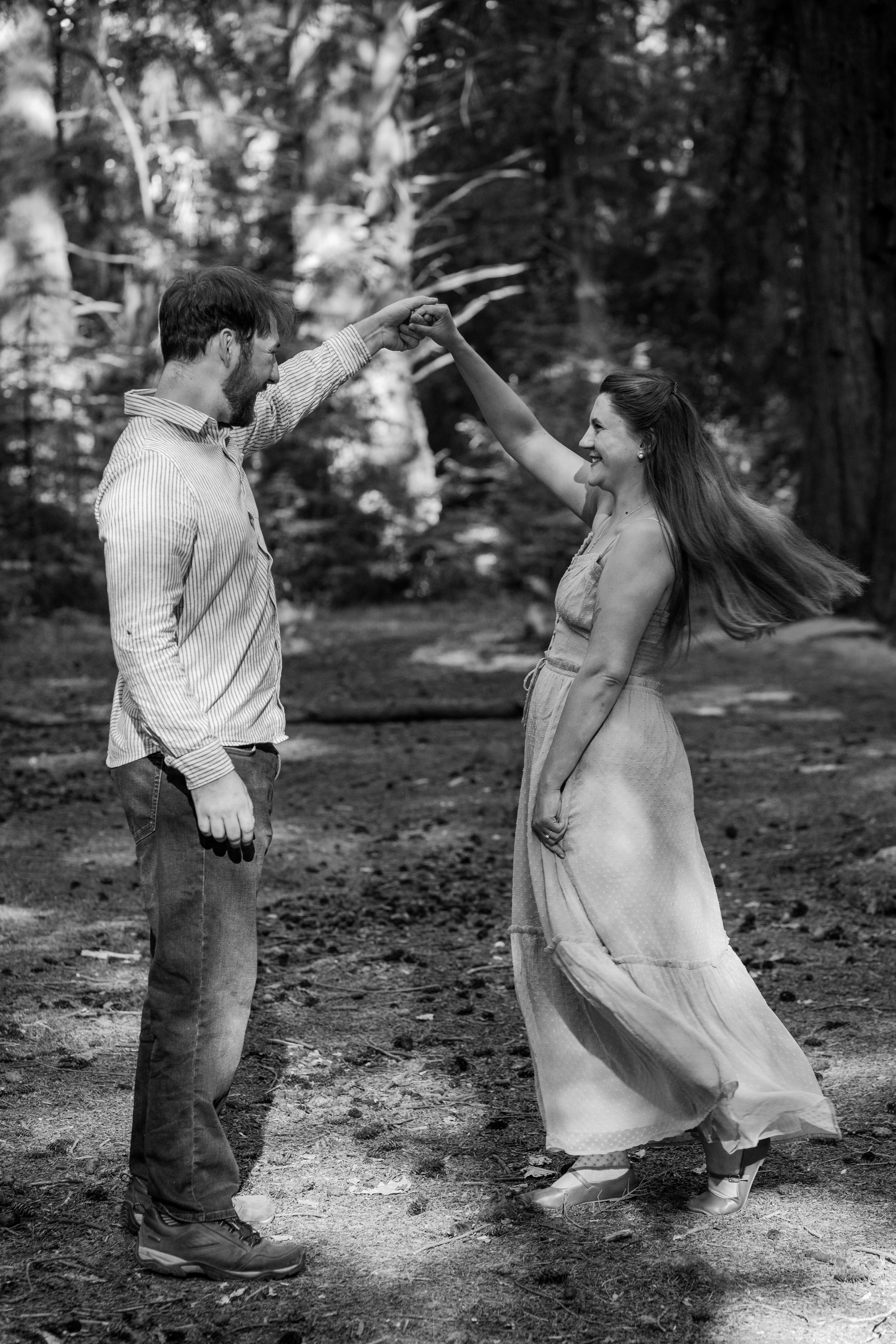 A black and white photo of a couple dancing outdoors in a wooded area. The man is wearing a striped shirt and jeans, and the woman is wearing a long dress with a full skirt. They are holding hands, smiling, and looking at each other.