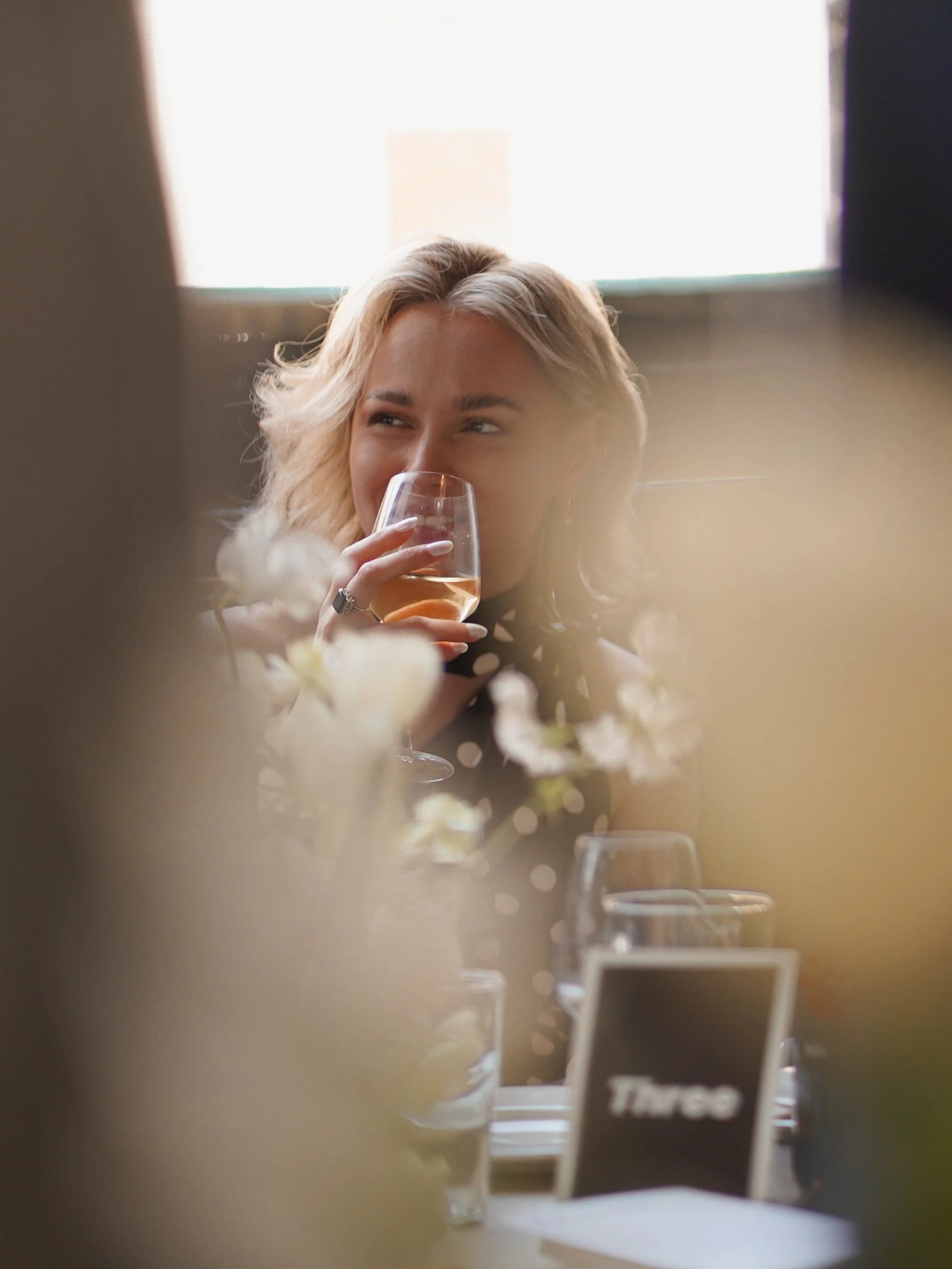 A woman with blonde hair holding a glass of wine, smiling and looking to the side, in a warmly lit setting.