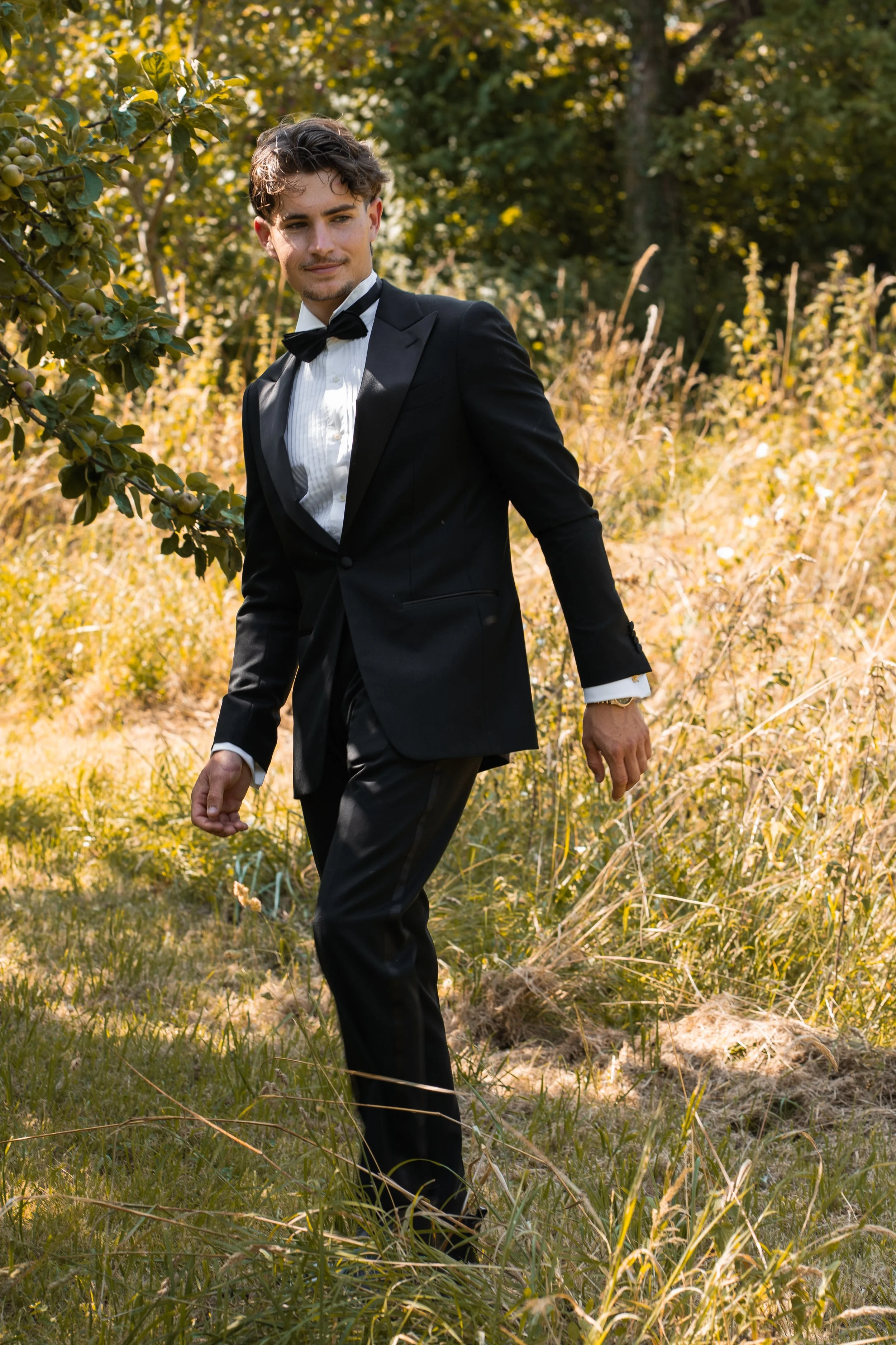 A young man dressed in a black tuxedo with a white shirt and black bow tie walking outdoors through a grassy and partly wooded area during daytime.