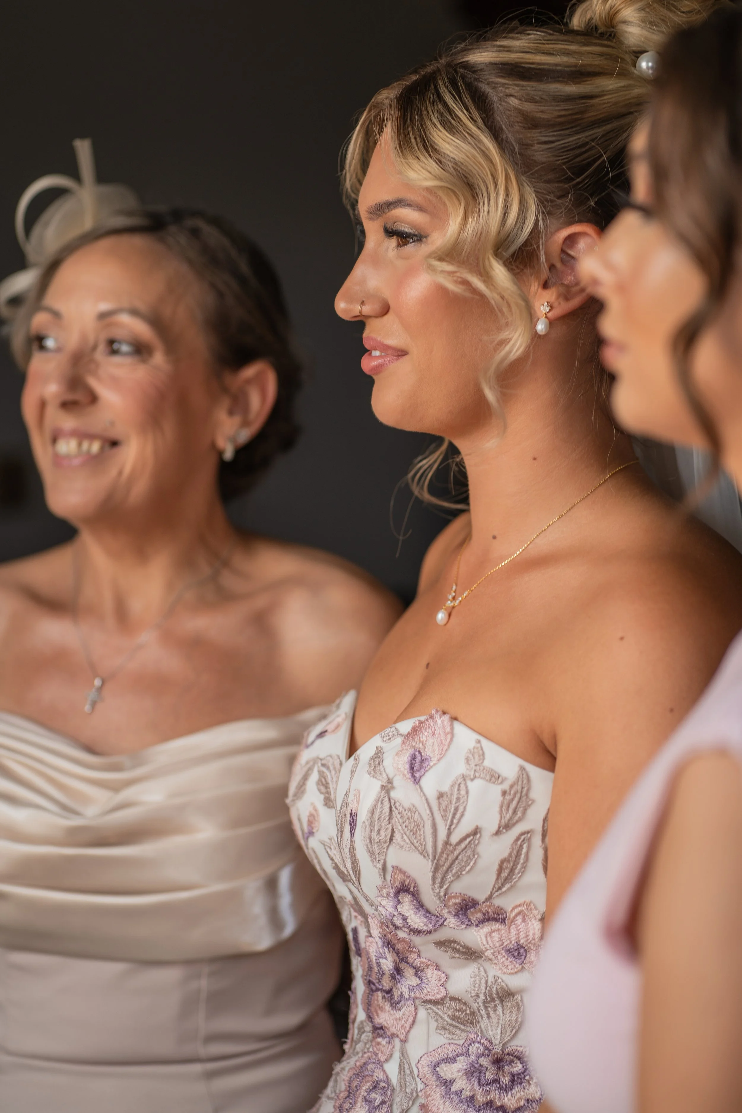 Close-up of three women in elegant dresses, wearing earrings and necklaces, with blurred background.