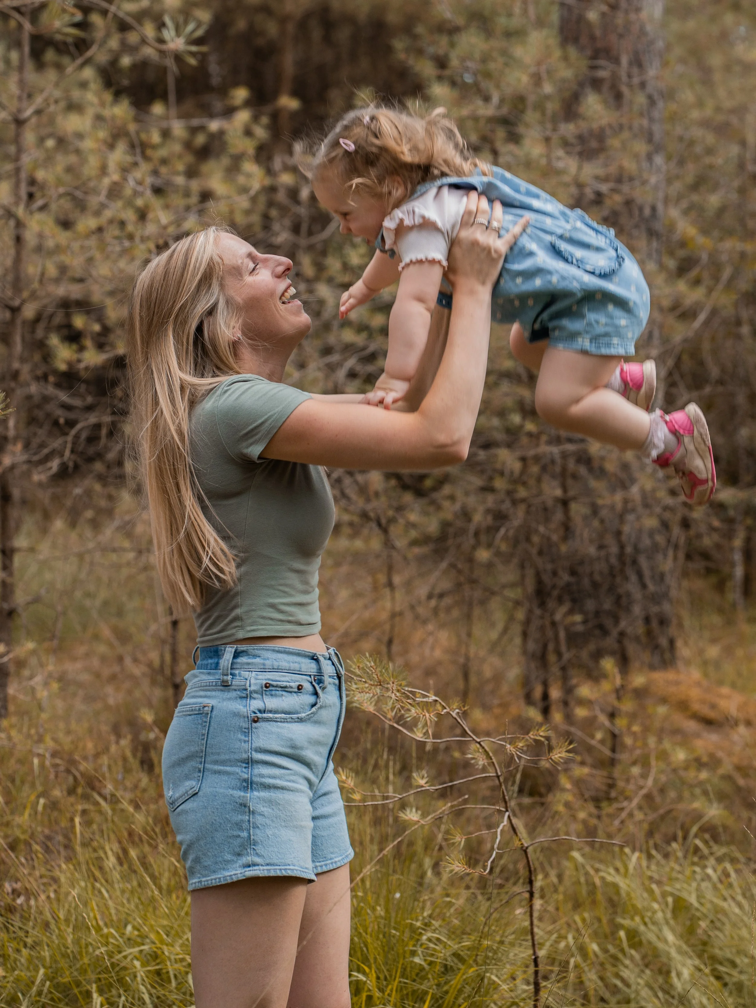 A woman with long blonde hair in a green T-shirt and denim shorts is holding a young girl with brown hair and pink bows, lifting her into the air outdoors surrounded by trees and tall grass.