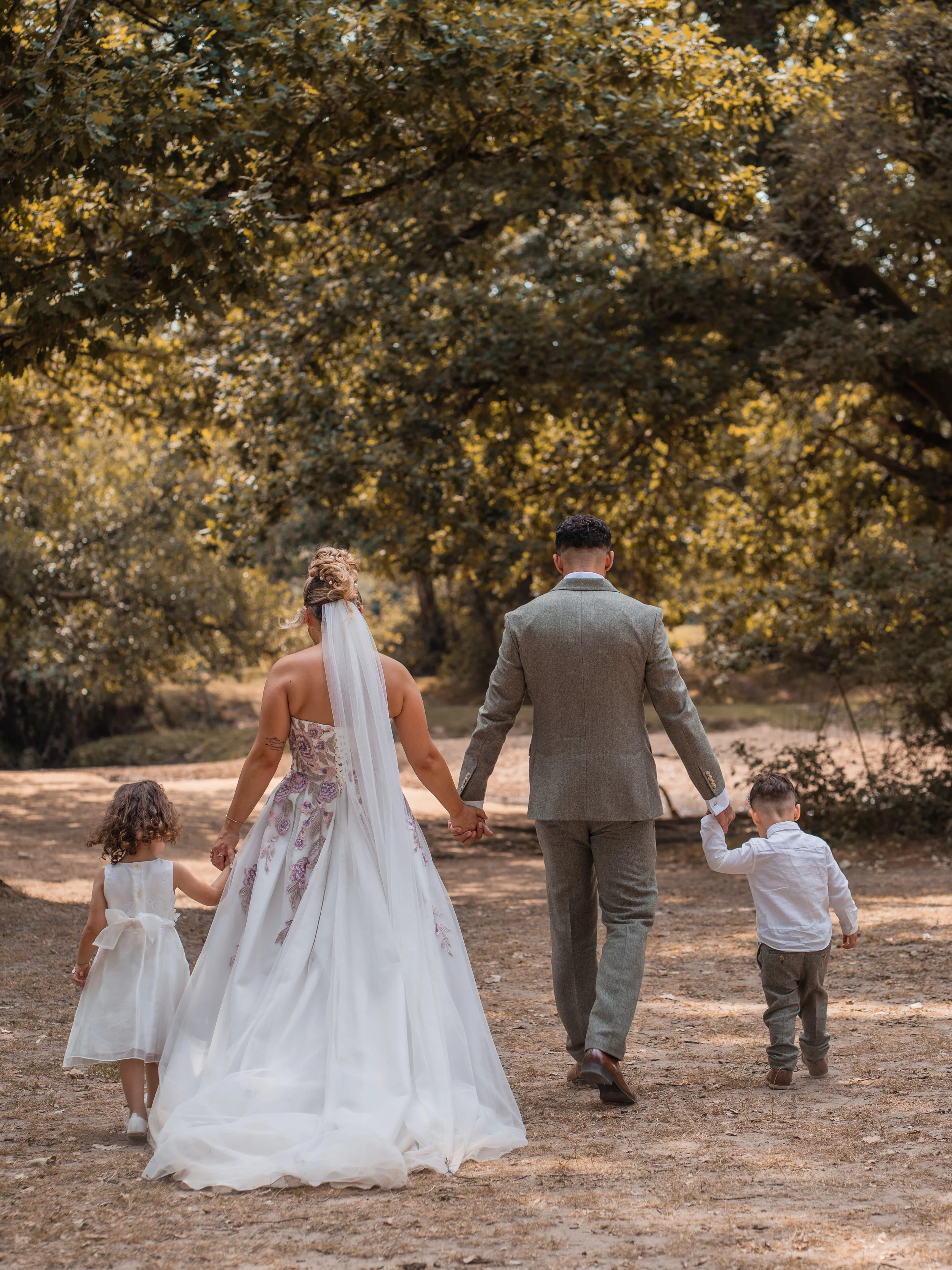 A couple, dressed in wedding attire, walking hand in hand outdoors with two children in white dresses and a young boy in a white shirt, under a large tree with yellow leaves, during daytime.