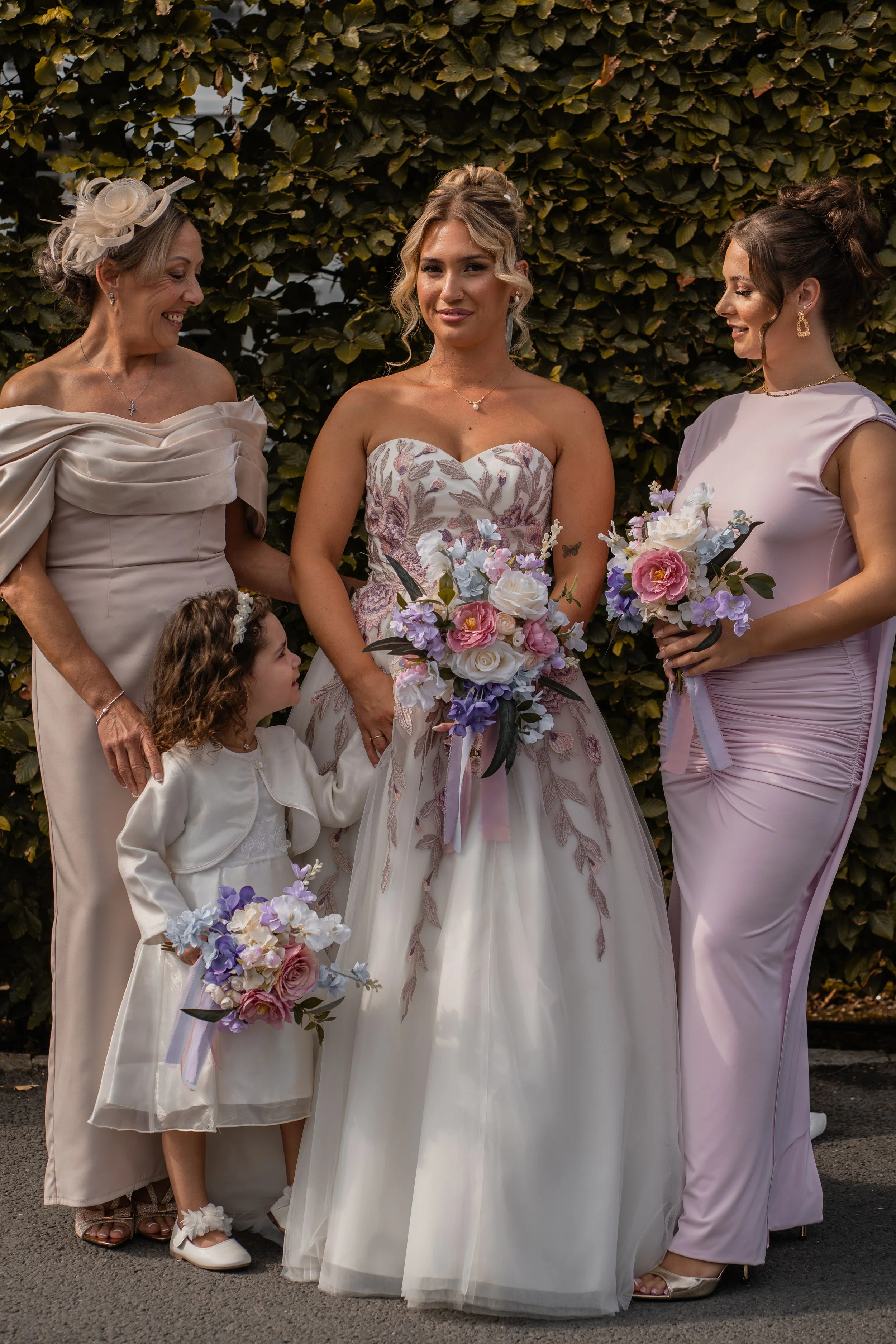 Four women and two young girls standing outdoors, dressed in formal attire, holding bouquets of flowers, with a hedge in the background. One woman in the center wears a strapless wedding gown, suggesting a wedding celebration.