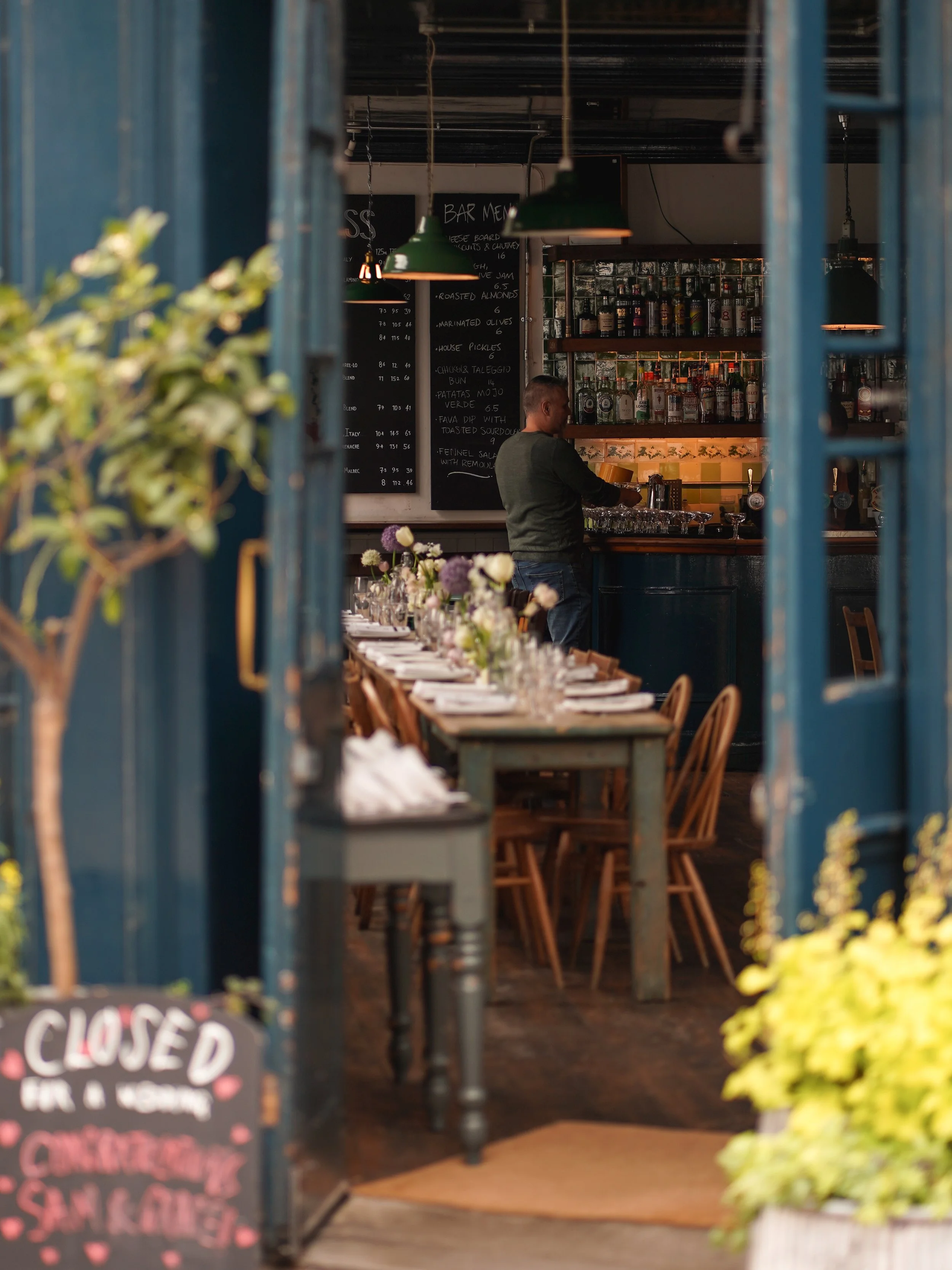 View of a cozy bar interior through an open door, featuring a man at the bar, chalkboard menu, shelves with bottles, and tables with floral centerpieces.