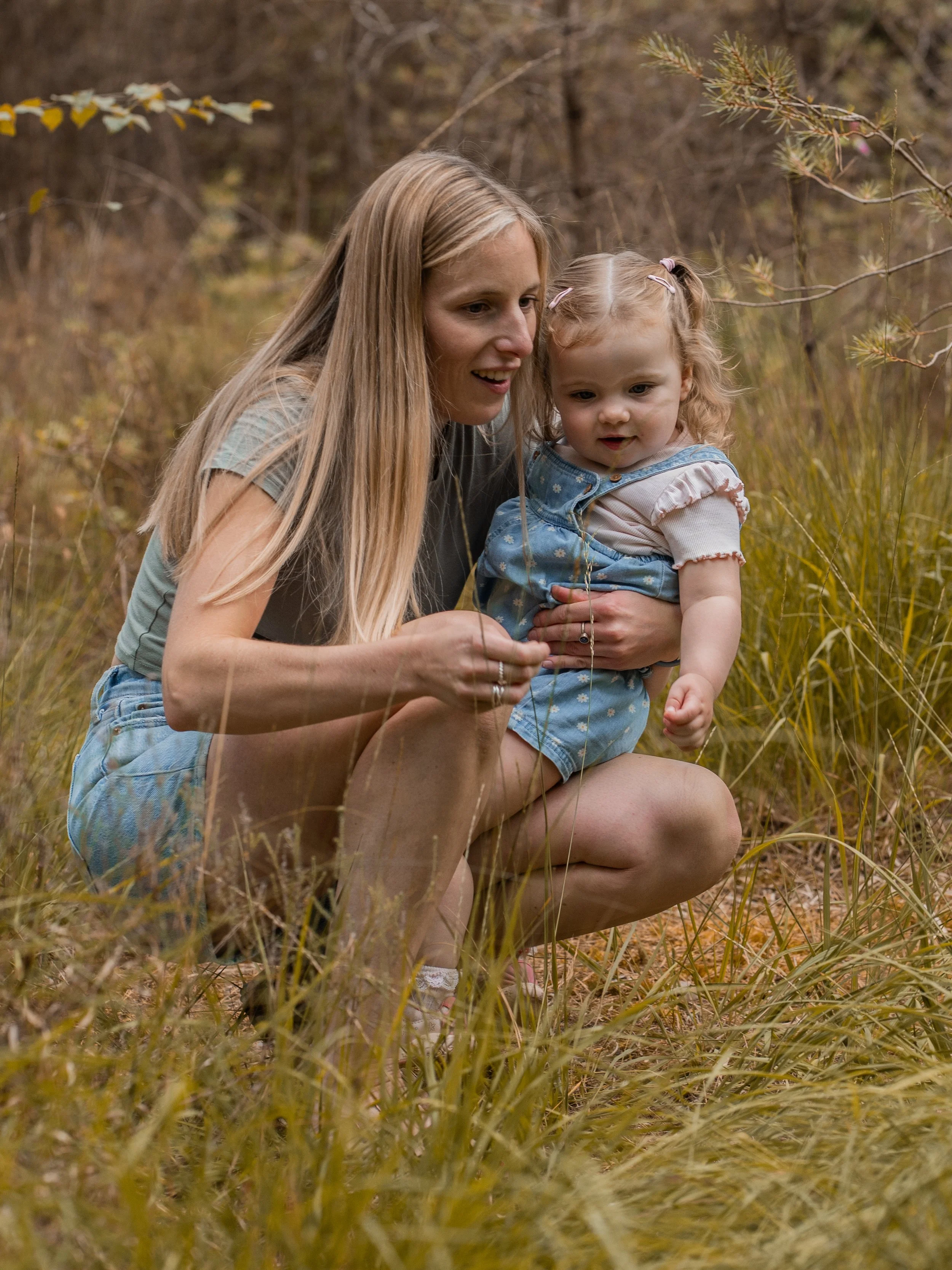 A woman with long blonde hair and a young girl with curly hair, hair clips, and a blue dress, sitting in tall grass in a wooded area, looking at something with interest.