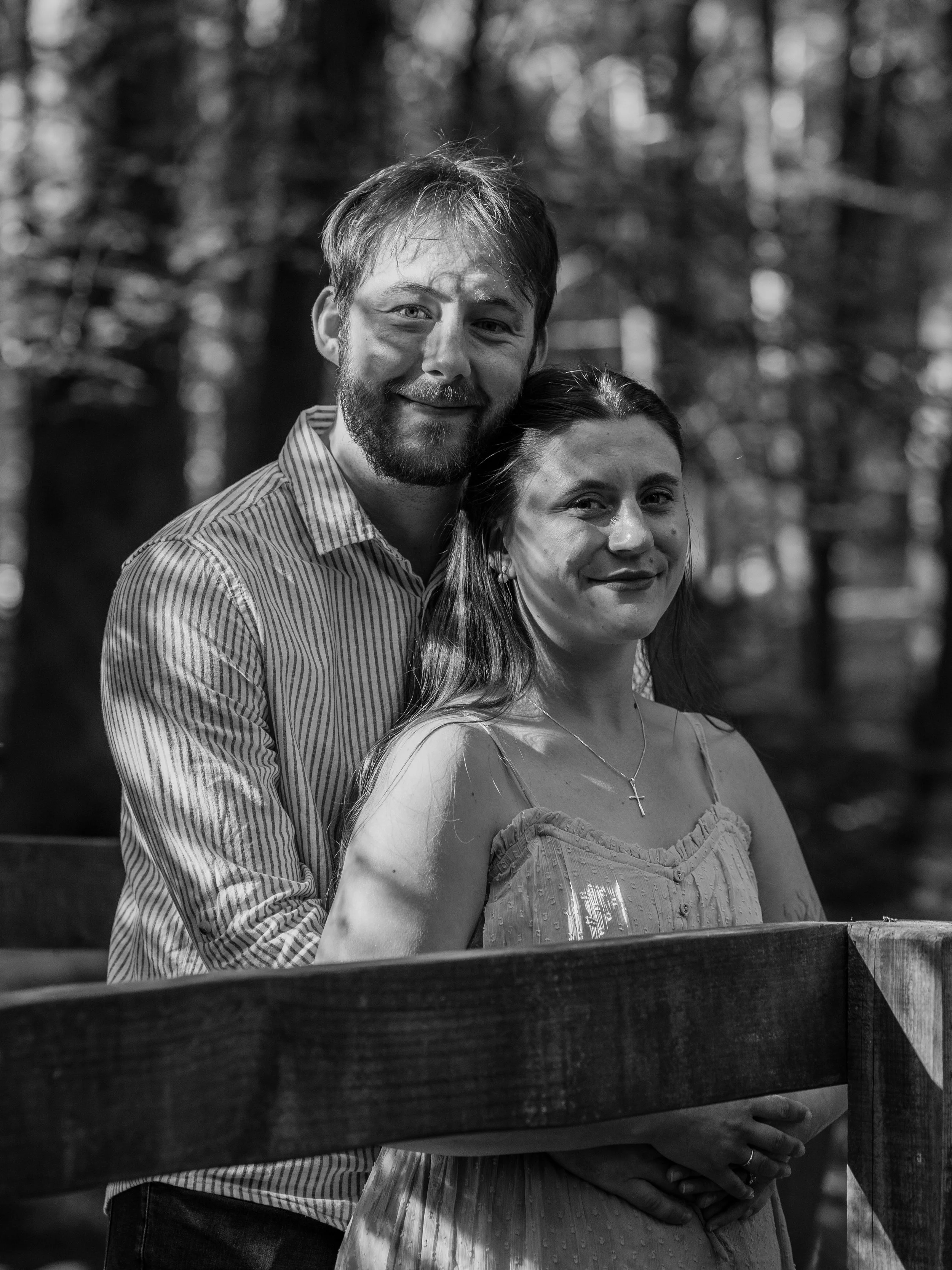 A black and white photograph of a smiling couple standing outdoors in front of a wooden fence, with a blurred background of trees. The man has a beard, mustache, and short hair, wearing a striped shirt. The woman has long hair, a necklace with a cros