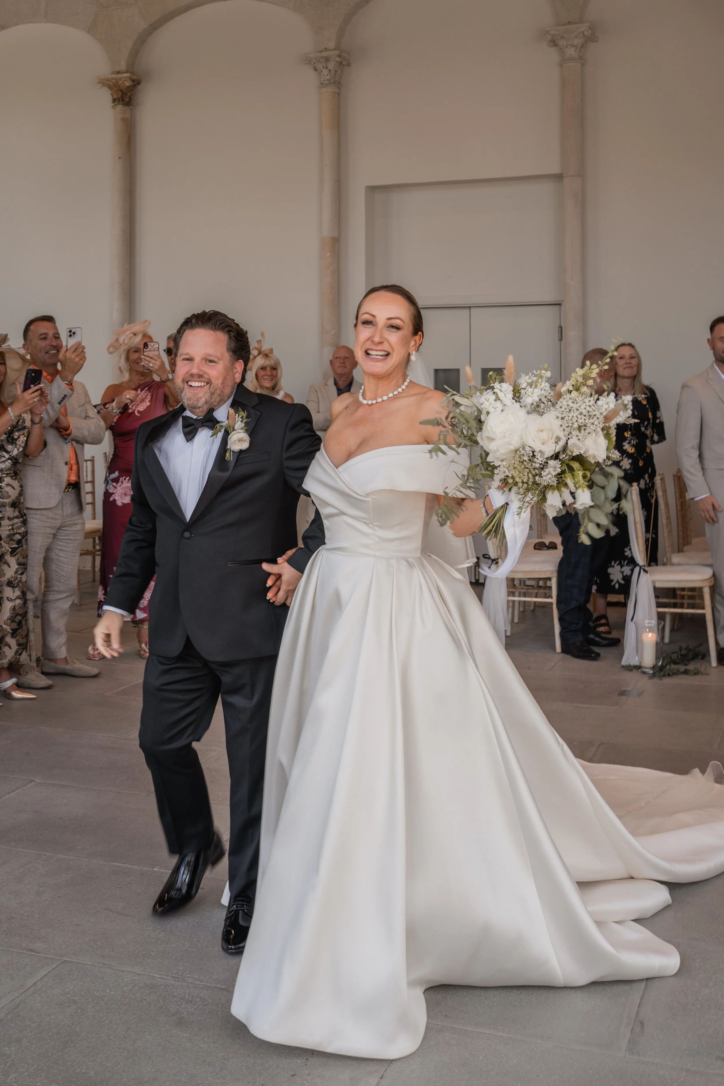 Bride and groom walking down the aisle during their wedding ceremony, surrounded by guests. The bride wears a white gown and holds a bouquet, while the groom wears a black tuxedo with a bow tie. Guests are smiling and taking photos.