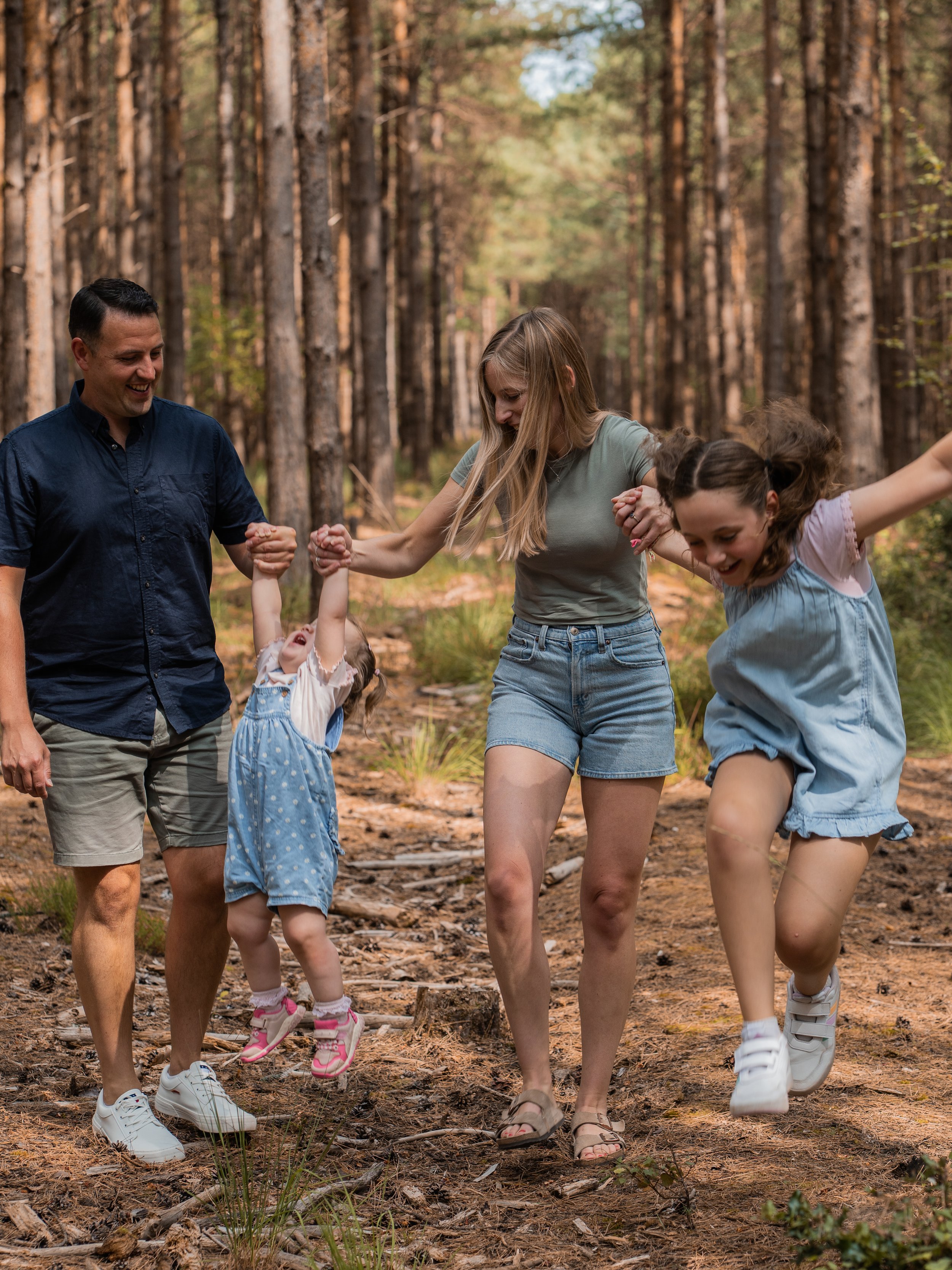 Family of four enjoying a walk in a pine forest, holding hands and smiling, with tall trees and sunlight filtering through the leaves.
