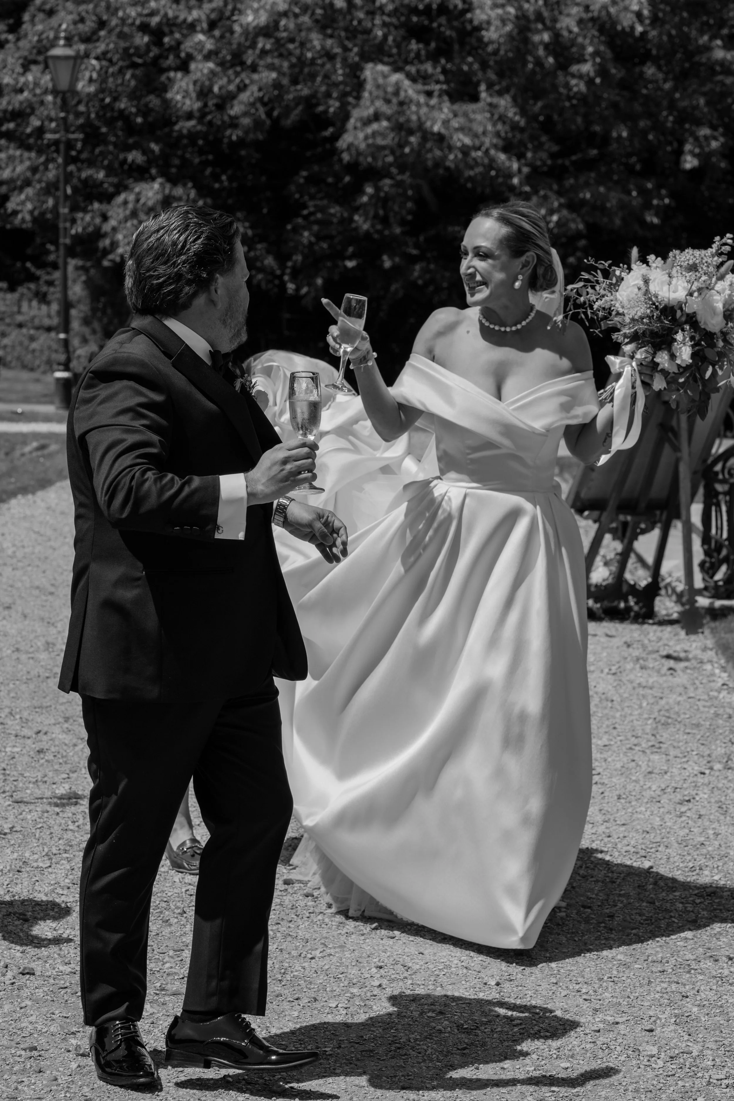 A woman in an off-the-shoulder wedding gown holding a bouquet and a man in a tuxedo holding a champagne flute, both smiling and dancing outdoors.