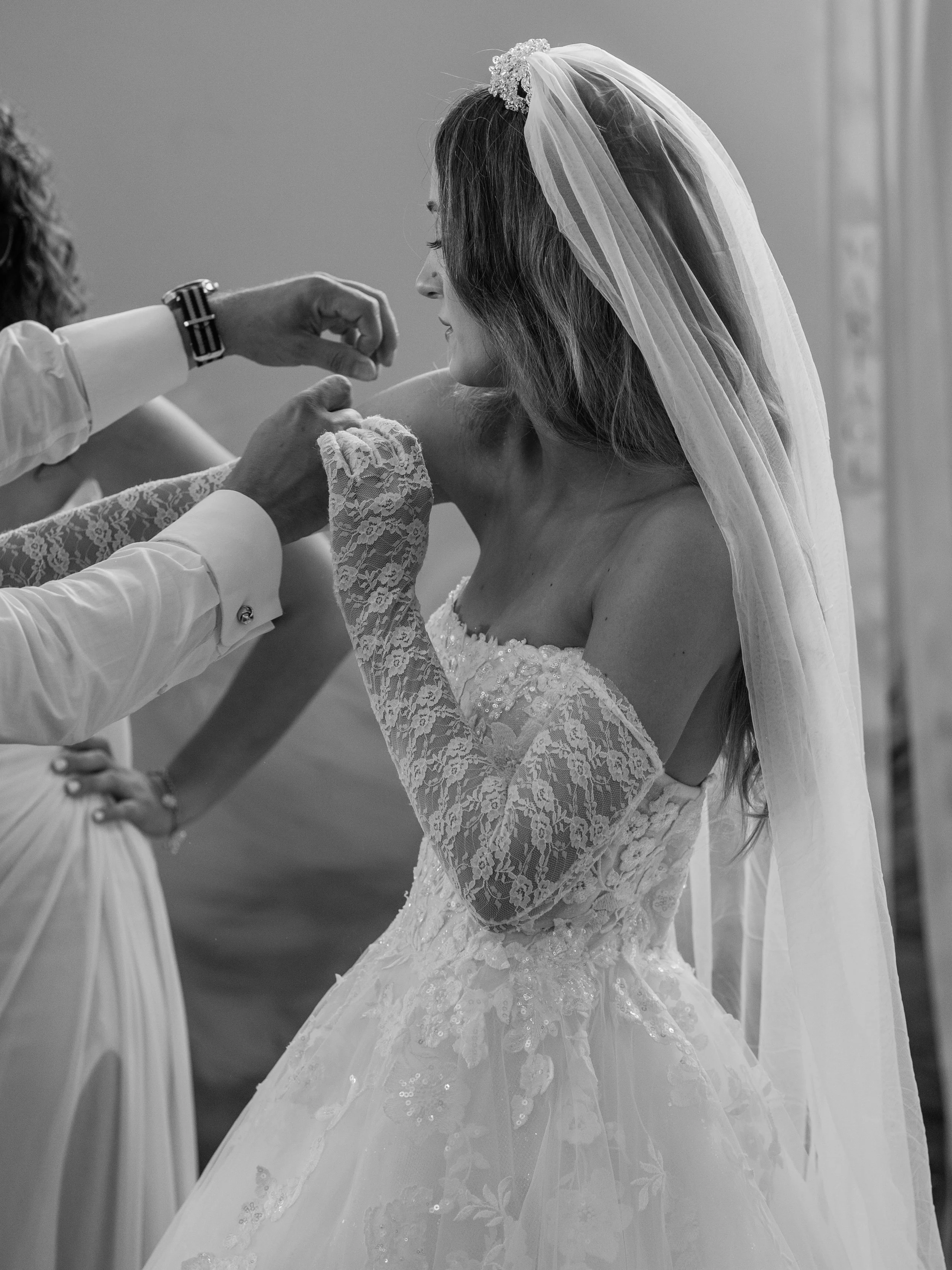 A bride in a wedding dress and veil is assisted by a person in a white shirt with a wristwatch, as they adjust her dress or gown, in black and white photograph.