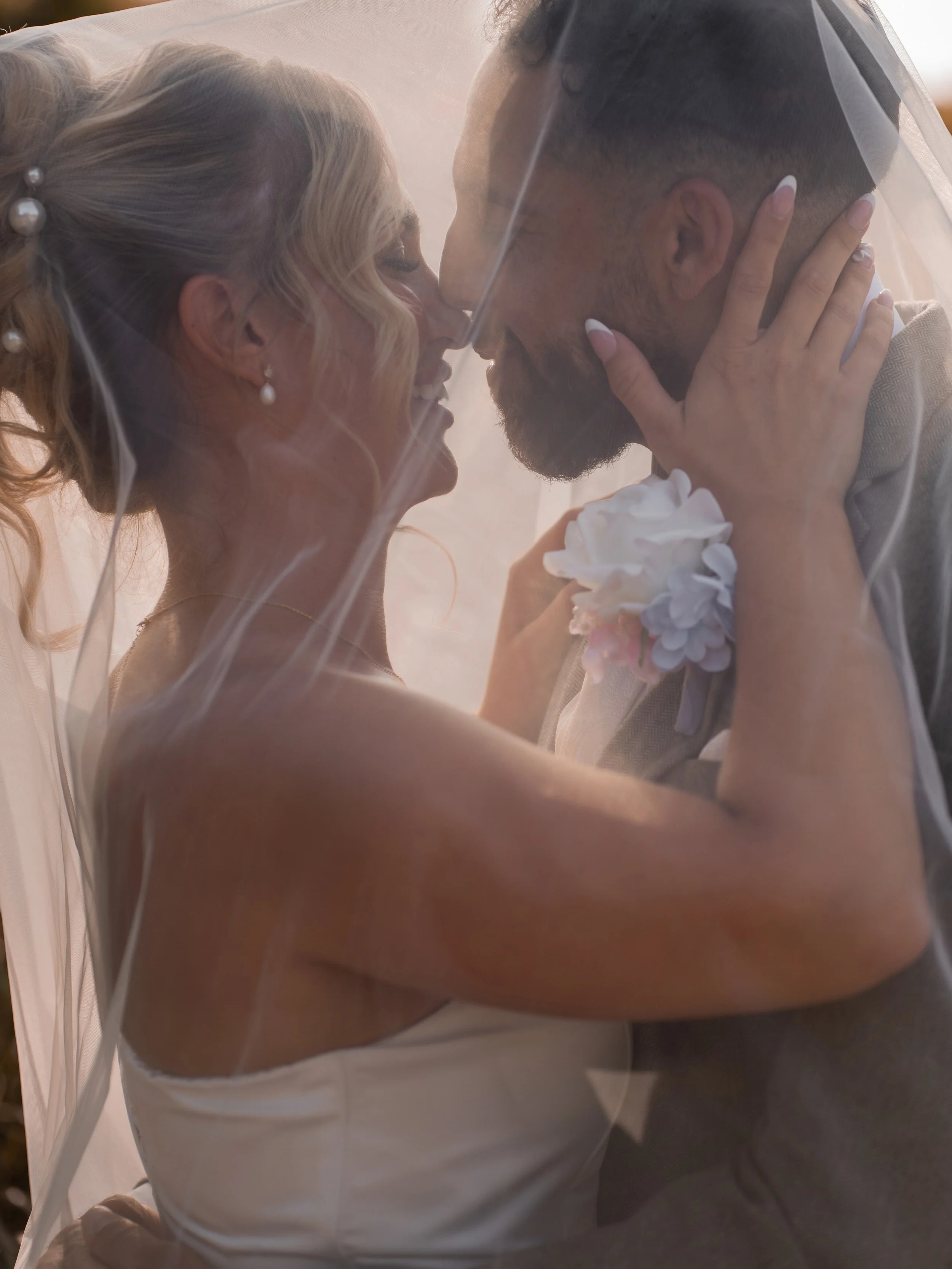 A bride and groom share a close moment with noses touching, beneath a veil, with the bride gently holding the groom's face during their wedding.