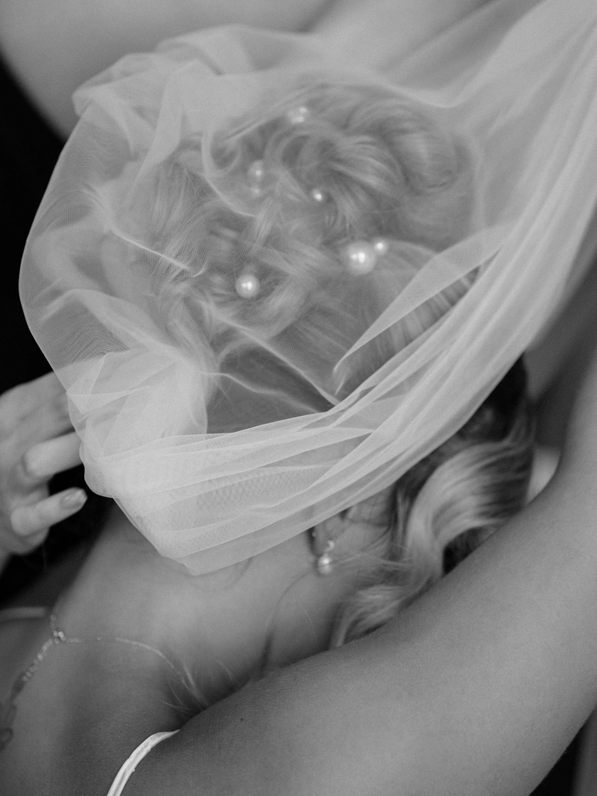 Close-up of a woman lying down with her face hidden under a sheer fabric, revealing her wavy hair, pearl necklace, and a tube top.