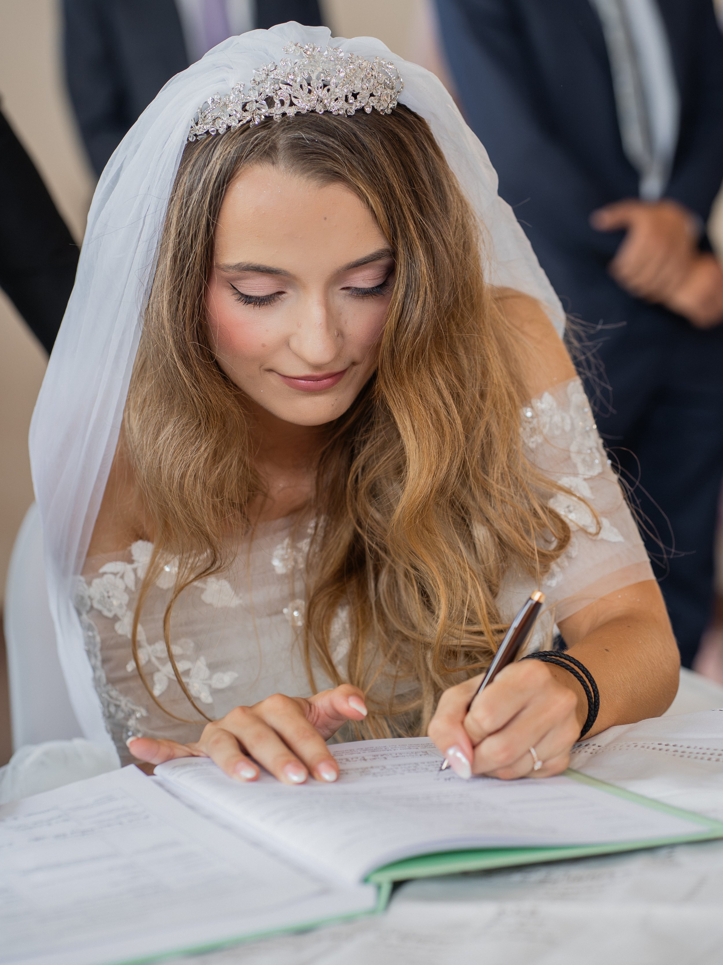 A woman in a wedding dress and tiara is signing a document with a pen, sitting at a table.