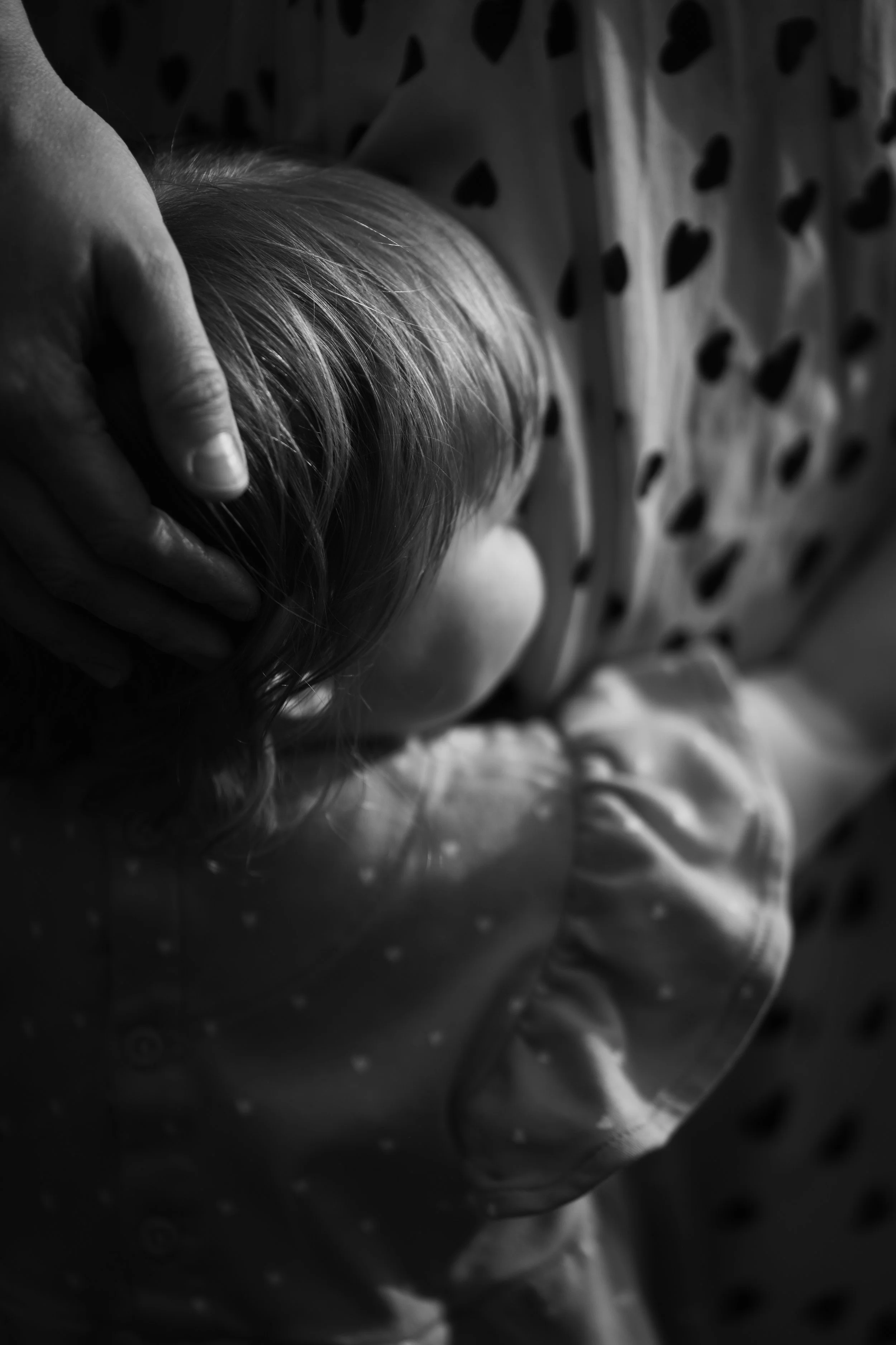 A close-up black and white photo of a person with short hair resting their head on a surface, with a hand gently holding their head. The person is wearing a garment with a heart pattern.