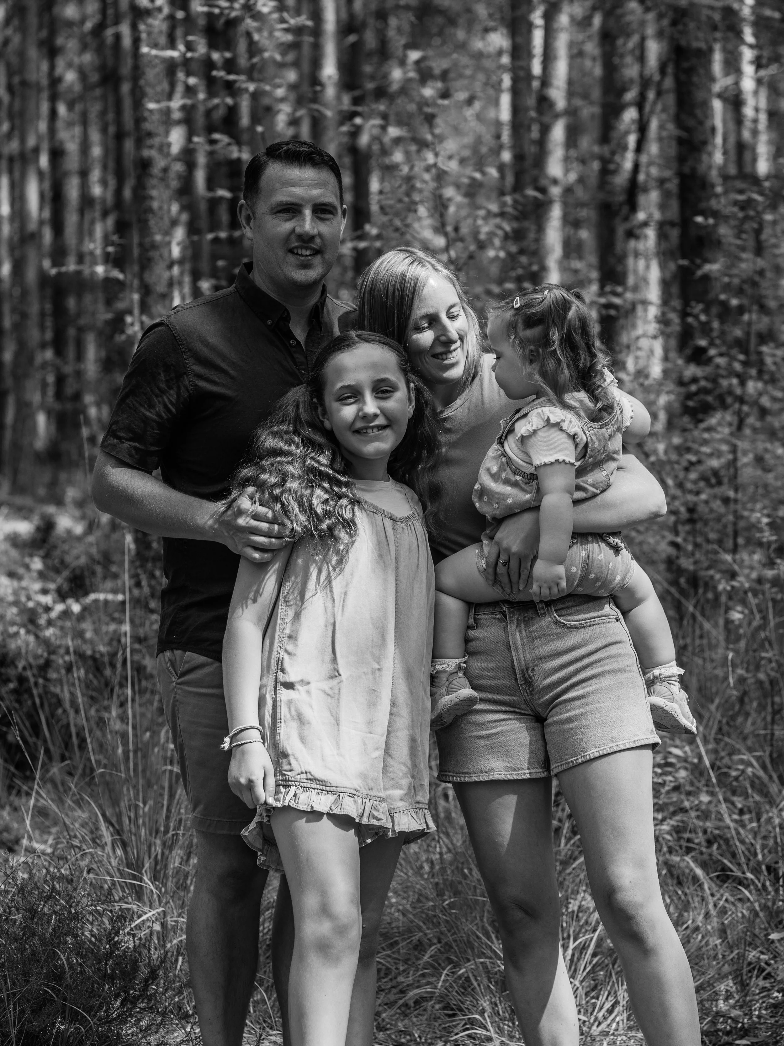 A black-and-white photograph of a family of four standing outdoors in a wooded area. The father, mother, and two young girls are smiling and close together, with the mother holding one of the girls.