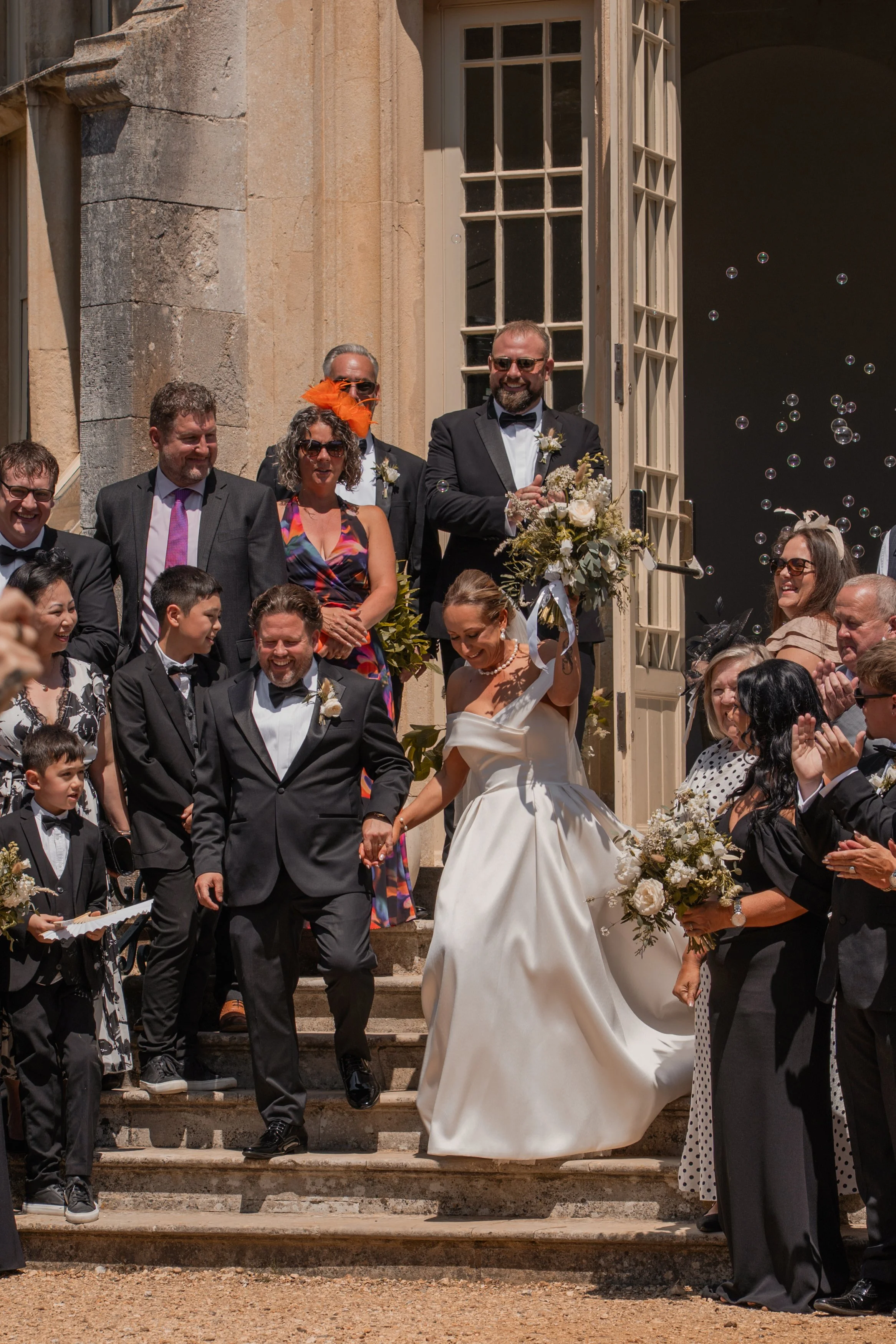 A wedding celebration on the steps of a historic building with multiple people dressed in formal attire, including the bride in a white wedding gown holding a bouquet, and the groom in a tuxedo, surrounded by friends and family.