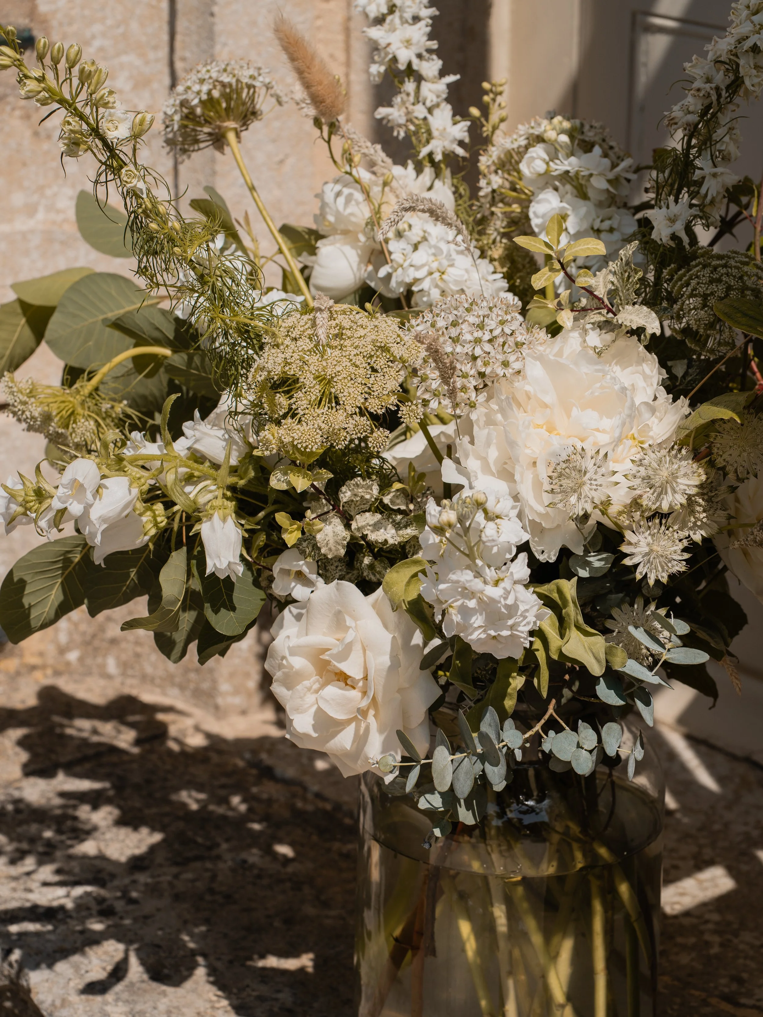 A bouquet of white flowers in a glass vase on a stone surface with sunlight casting shadows.