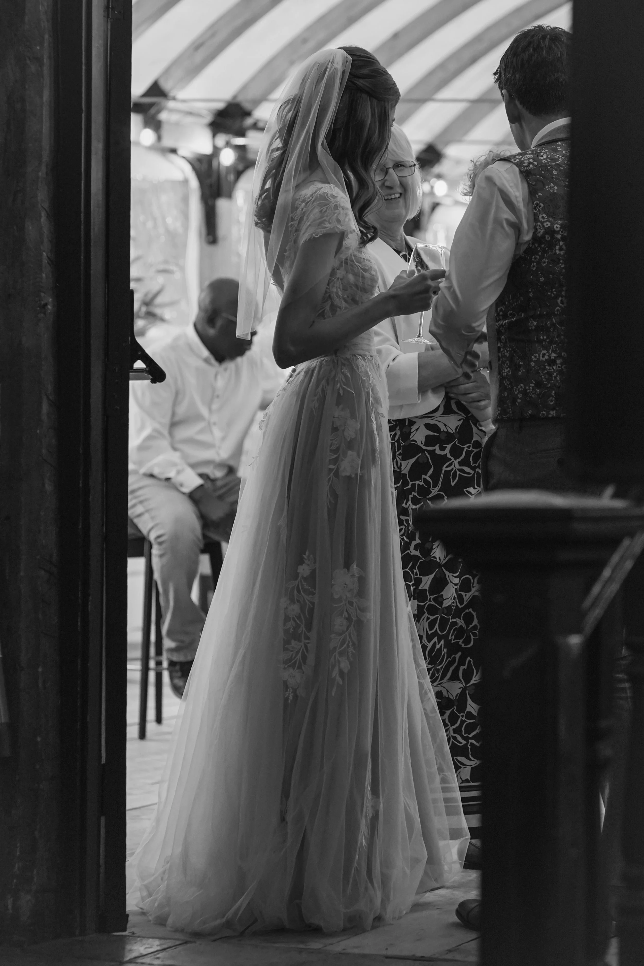 A black and white photo of a bride in a wedding dress and veil holding a champagne flute, smiling and talking to a man. A woman with glasses stands nearby, also smiling. In the background, a seated man appears to be looking away.