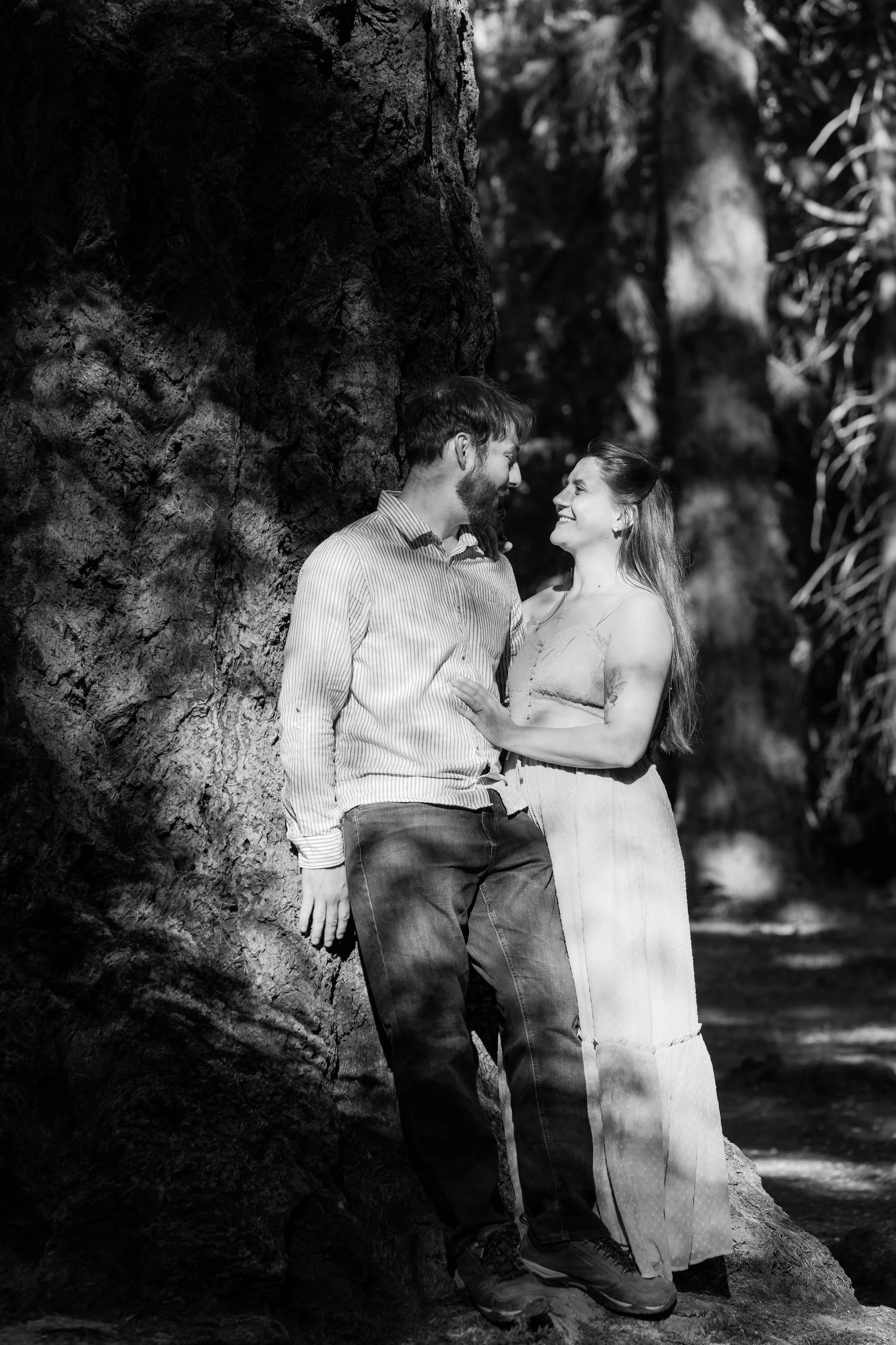 A black and white photo of a man and woman standing close together outdoors, smiling and looking at each other near a large tree.