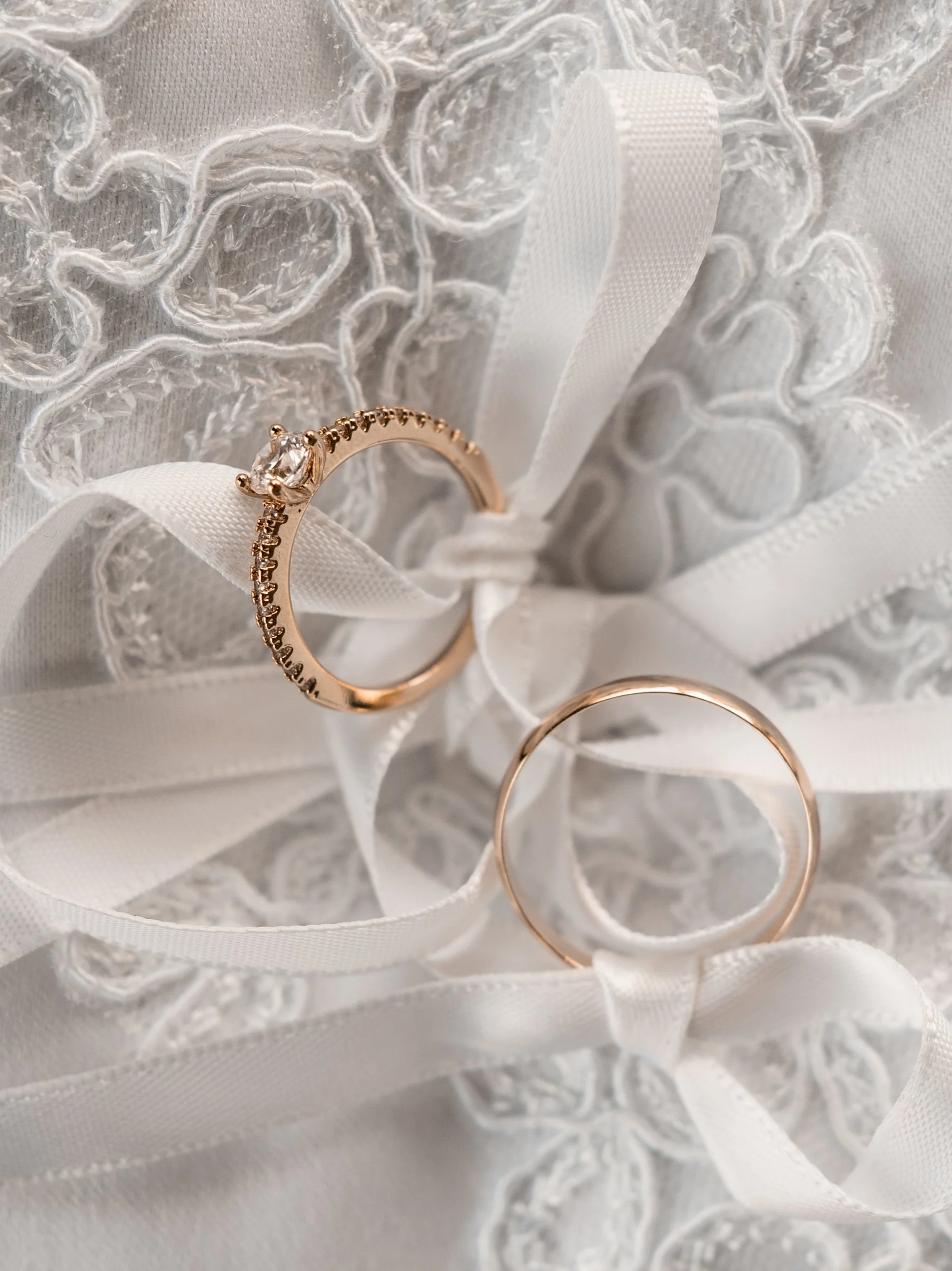 A close-up of a gold engagement ring with a central diamond, placed on white lace fabric with decorative embroidery, accompanied by a plain gold wedding band.