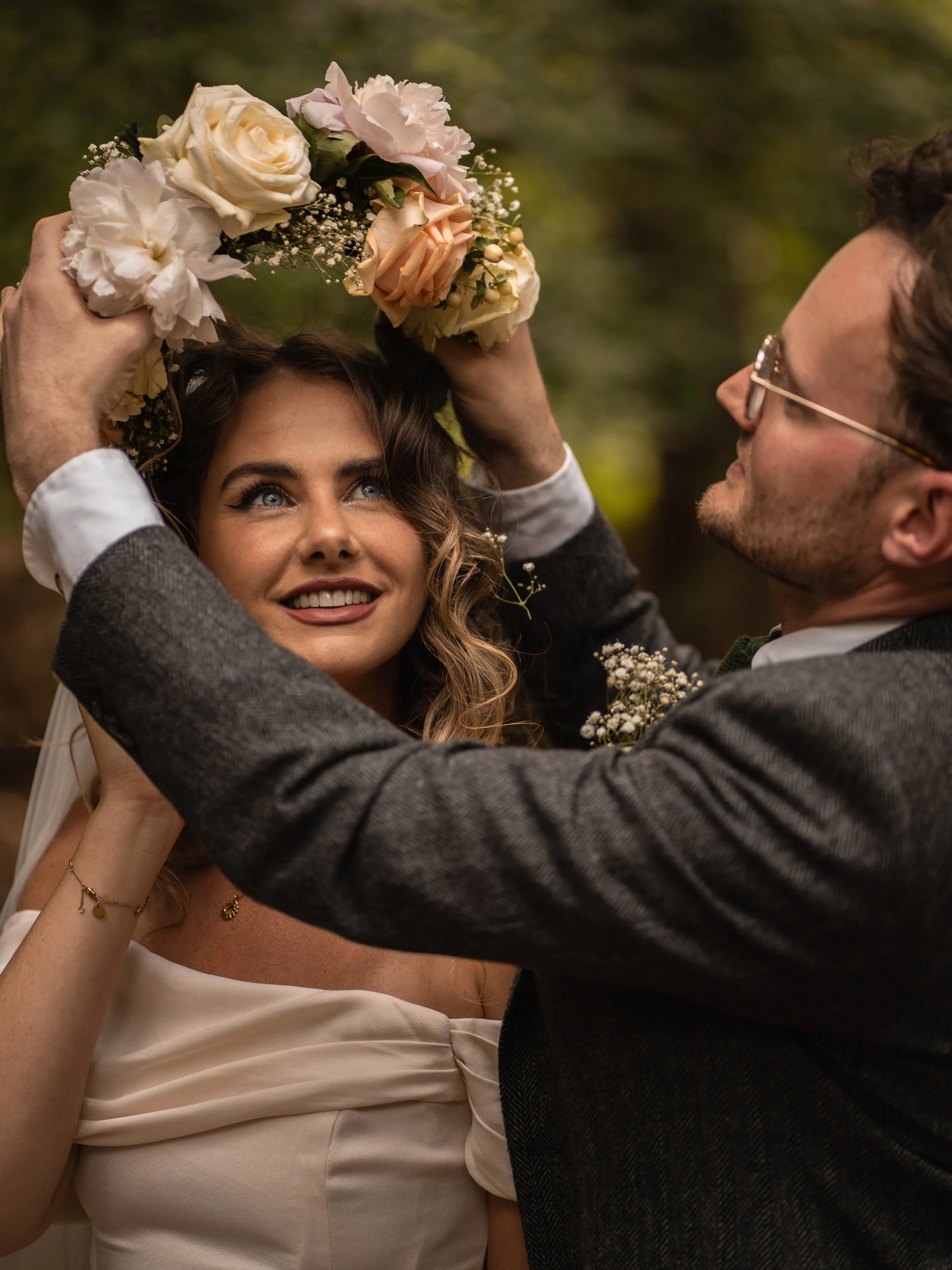 A man and woman at a wedding, with the man placing a floral crown on the woman's head. The woman has wavy brown hair, bright blue eyes, and is smiling. She is wearing an off-white dress with a draped sleeve. The man has short curly hair, glasses, and