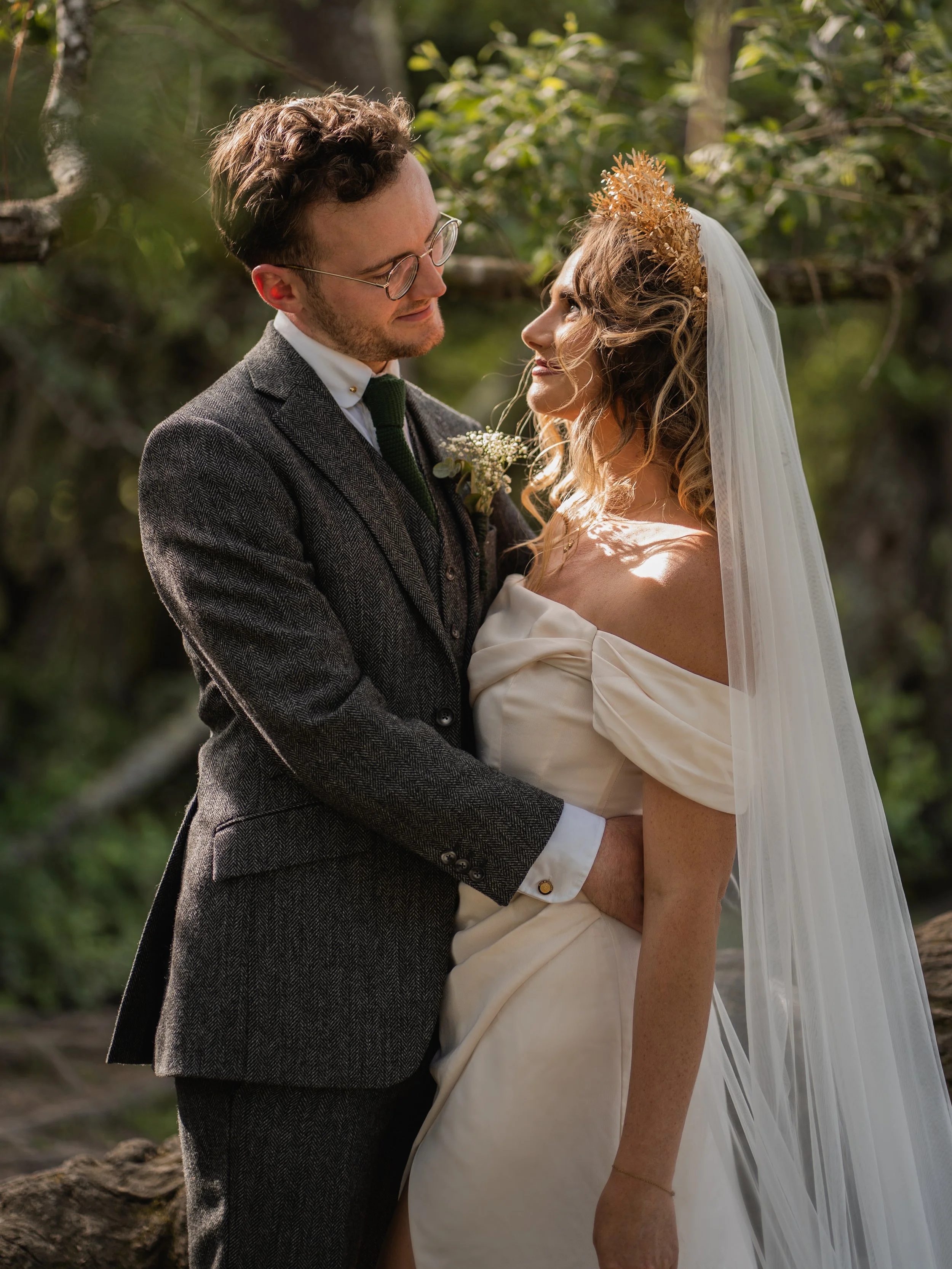 A bride and groom look into each other's eyes in a forest setting, dressed in wedding attire with natural light illuminating their faces.