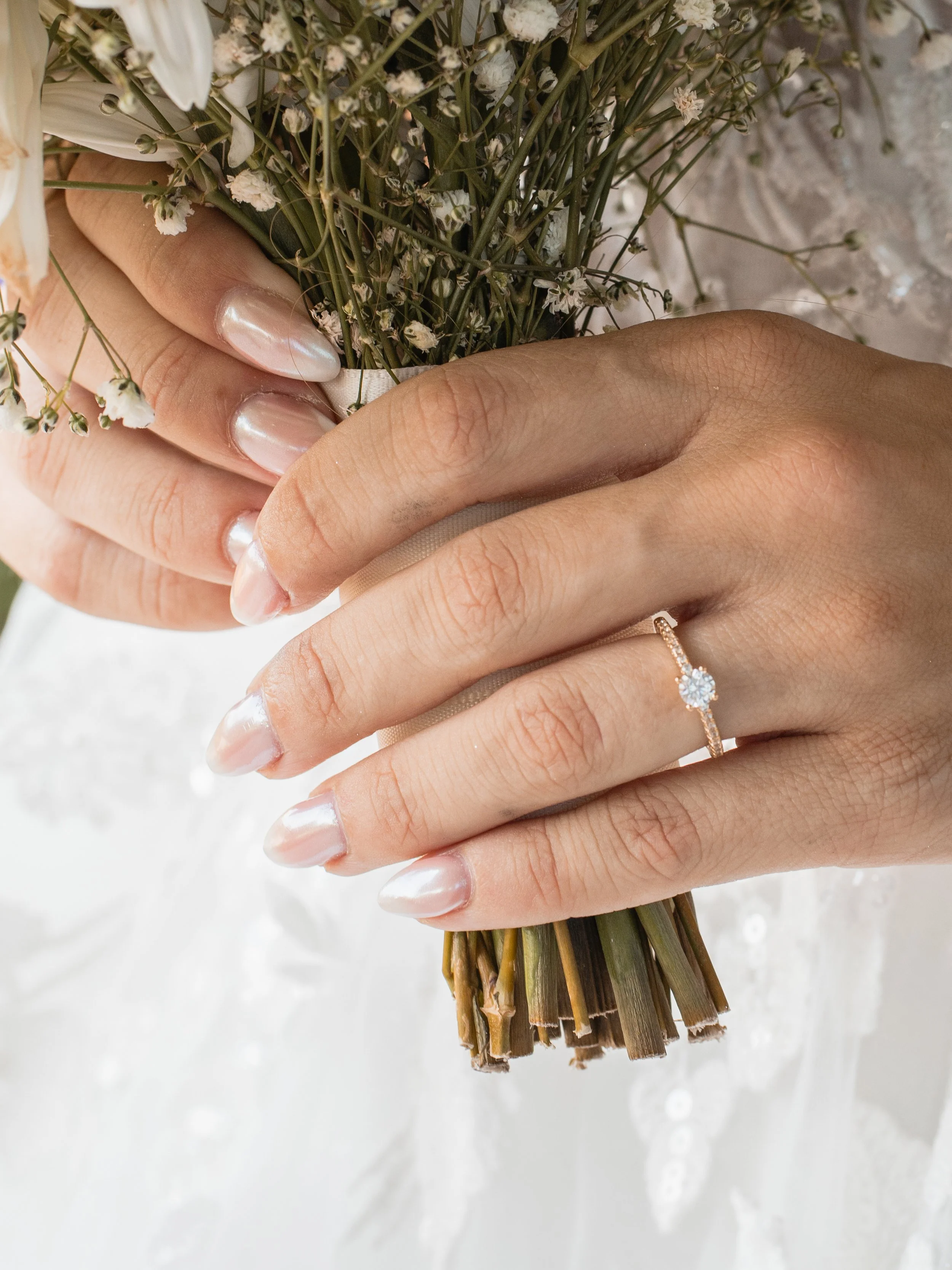 Close-up of a woman's hand with a diamond engagement ring, holding a bouquet of small white flowers and greenery.