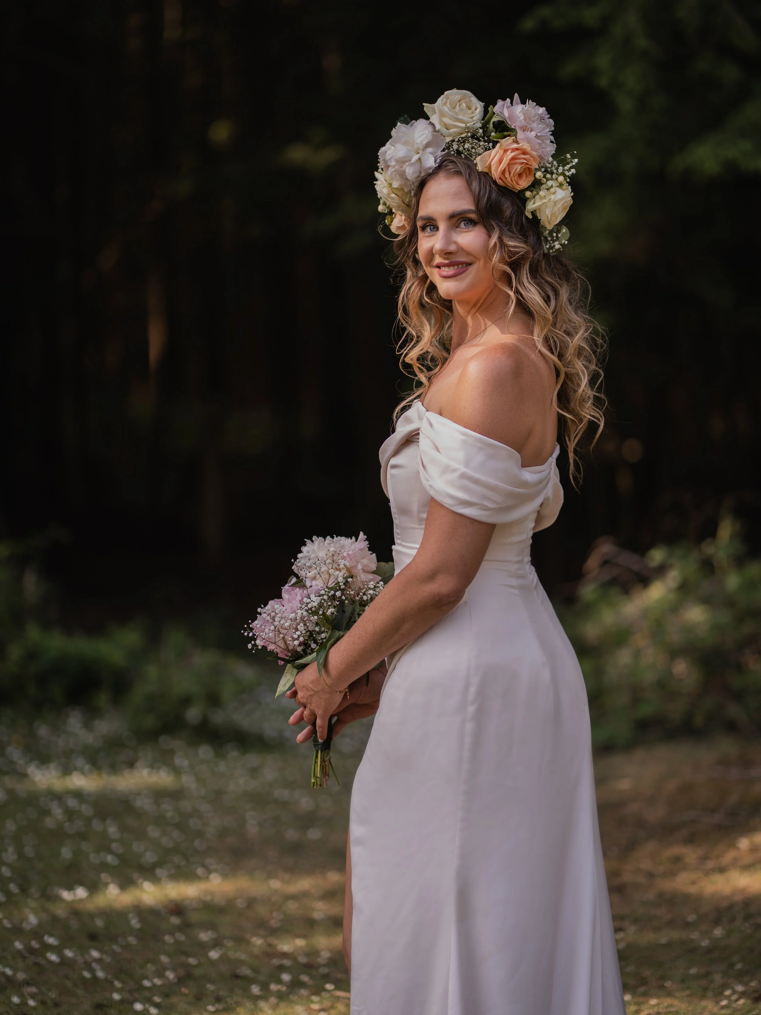 A woman in a white dress standing outdoors holding a bouquet of pink and white flowers, wearing a floral crown made of white, blush, and peach roses and other small white flowers, with a blurred dark forest background.