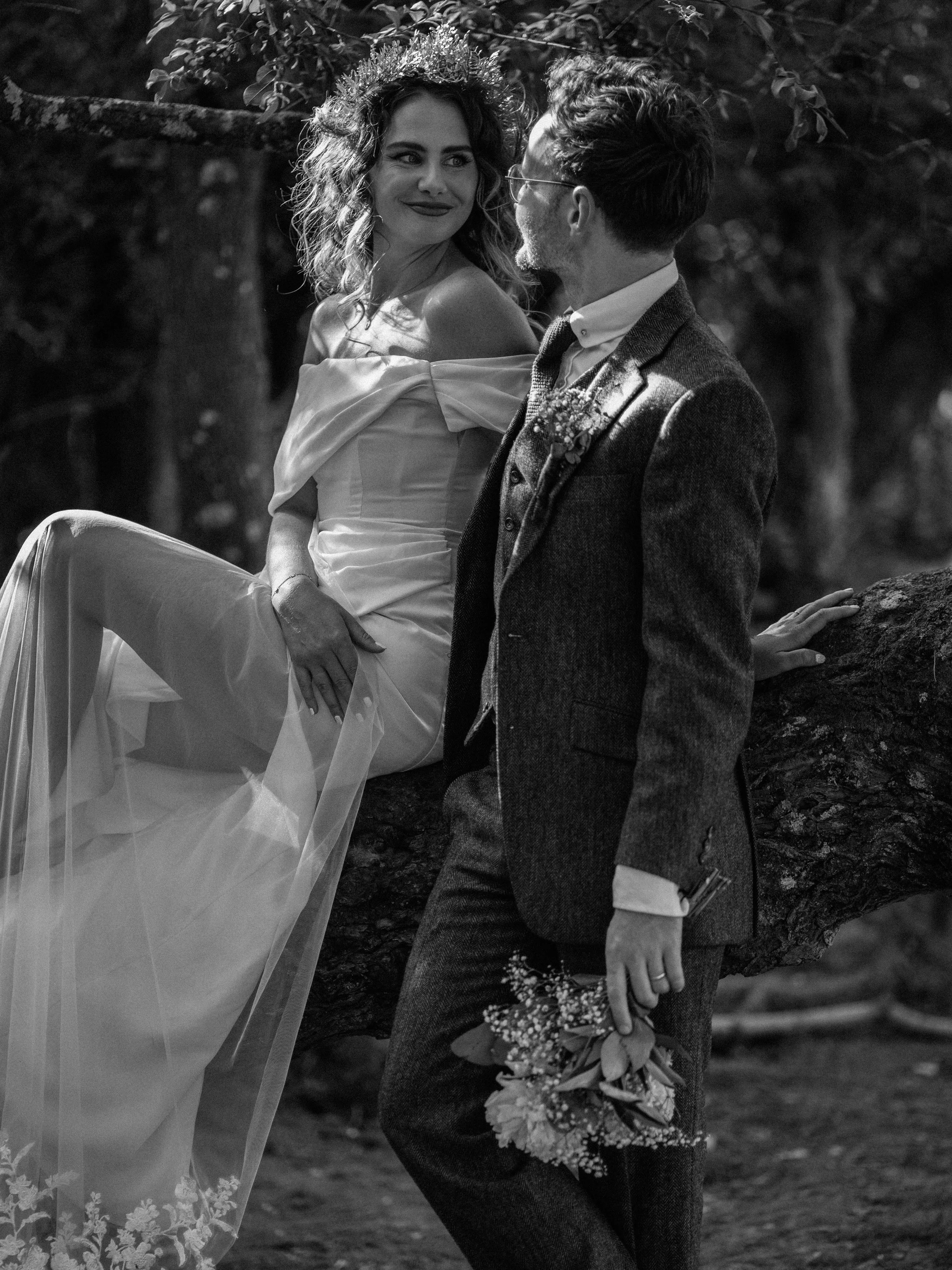 A black and white photo of a woman in a wedding dress and a crown, sitting on a tree branch, looking at a man in a tuxedo who is standing beside her and holding a bouquet of flowers, both smiling and sharing a moment outdoors.