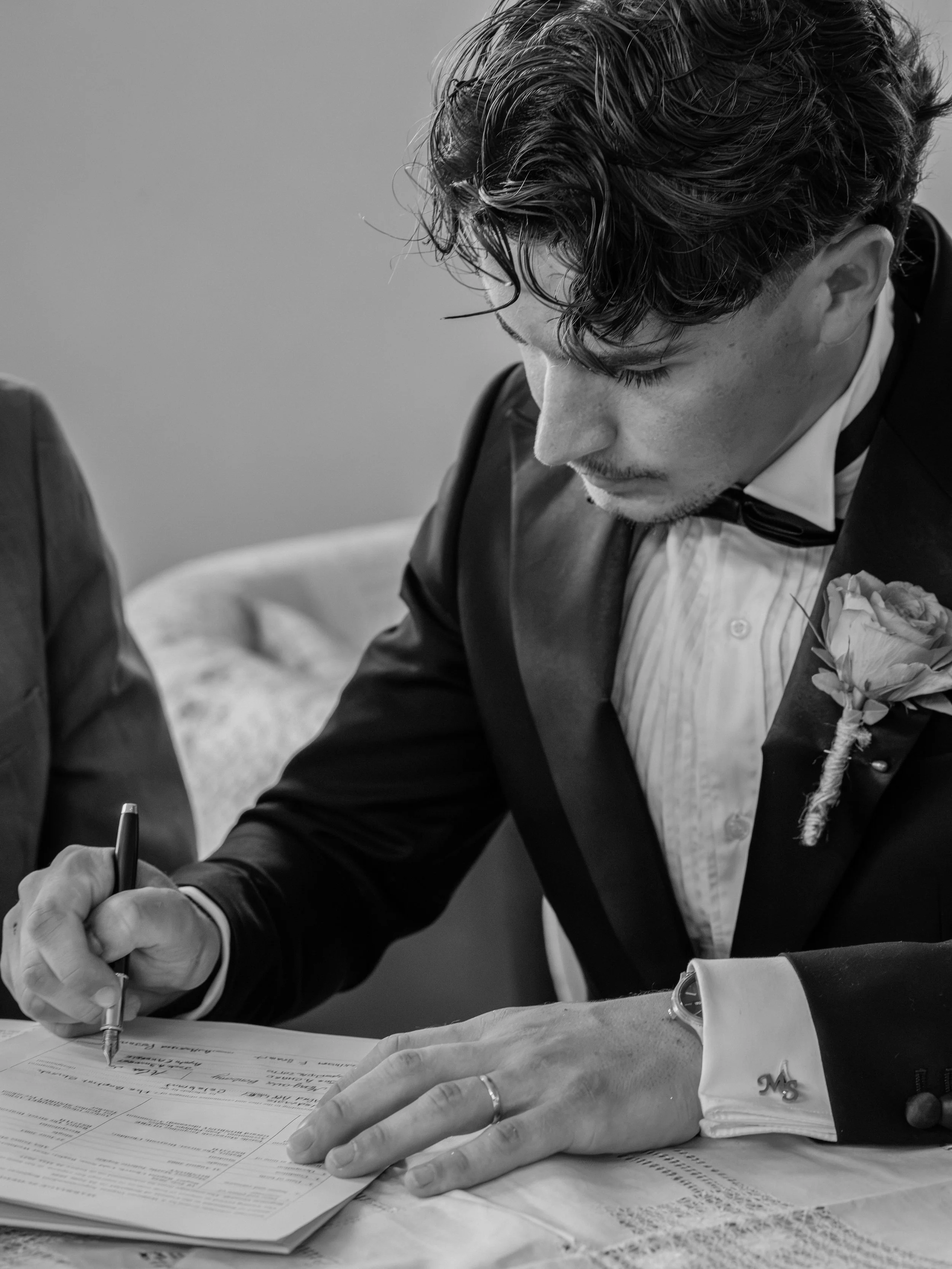 A man in a tuxedo with a bow tie and boutonniere signs a document at a table.