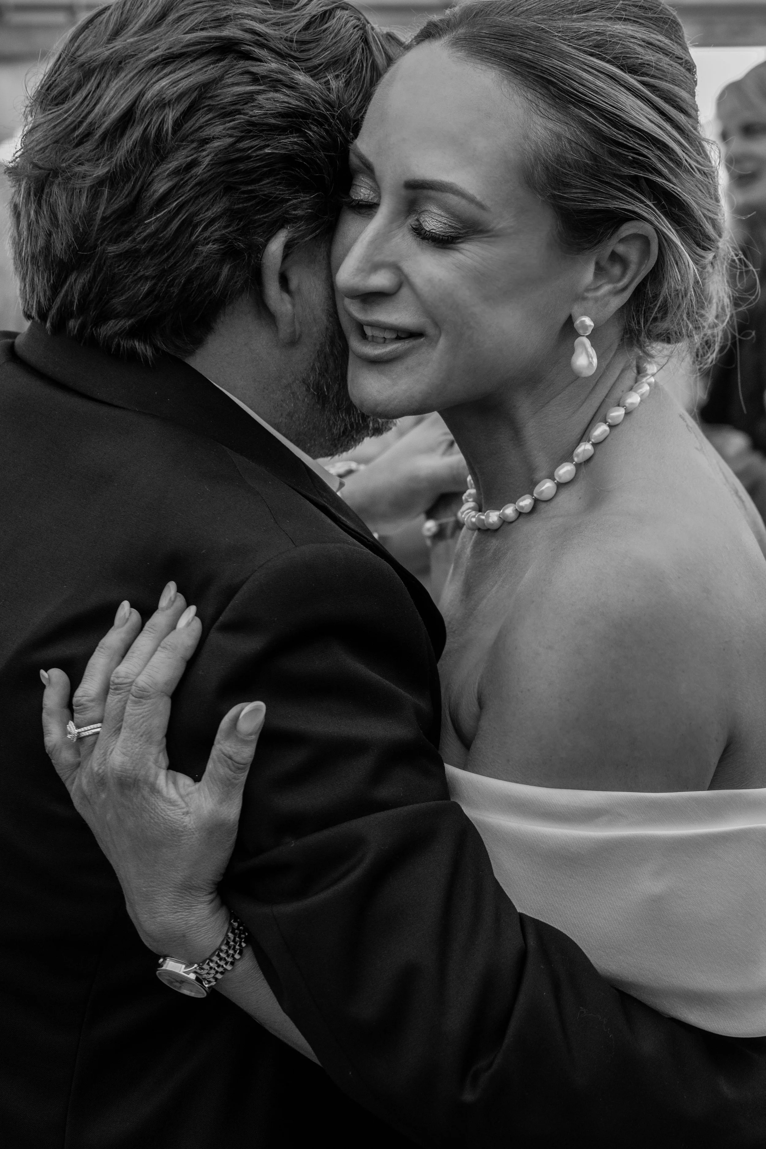 A black and white photo of a man and a woman hugging each other at a formal event. The man has dark hair and a beard, while the woman has short, styled hair and is wearing pearl jewelry. The woman's hand, showing a ring and a bracelet, is resting on the man's shoulder.