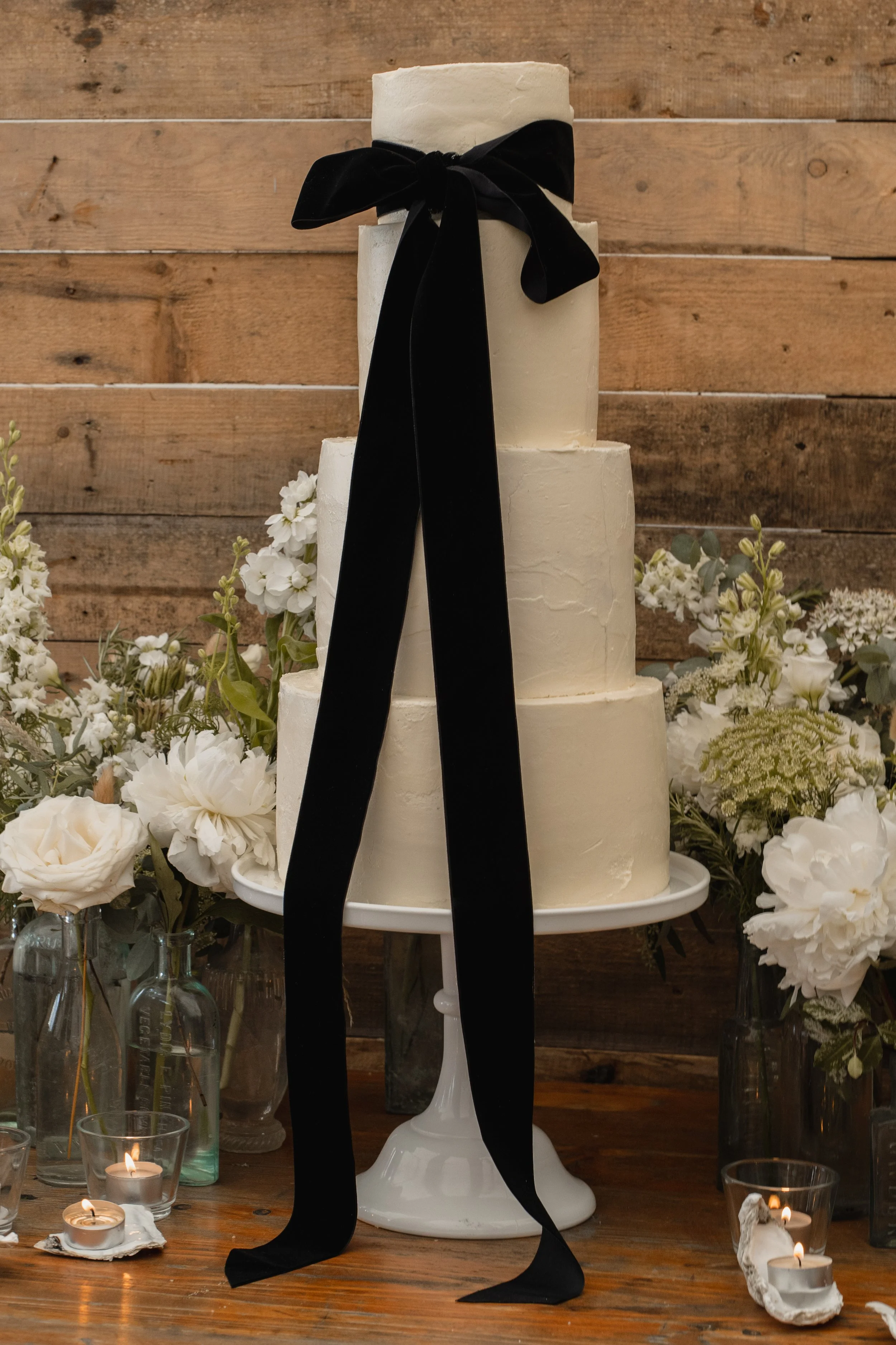 A three-tier white wedding cake with a black ribbon tied into a bow on top, surrounded by white flowers and candles, against a wooden wall background.