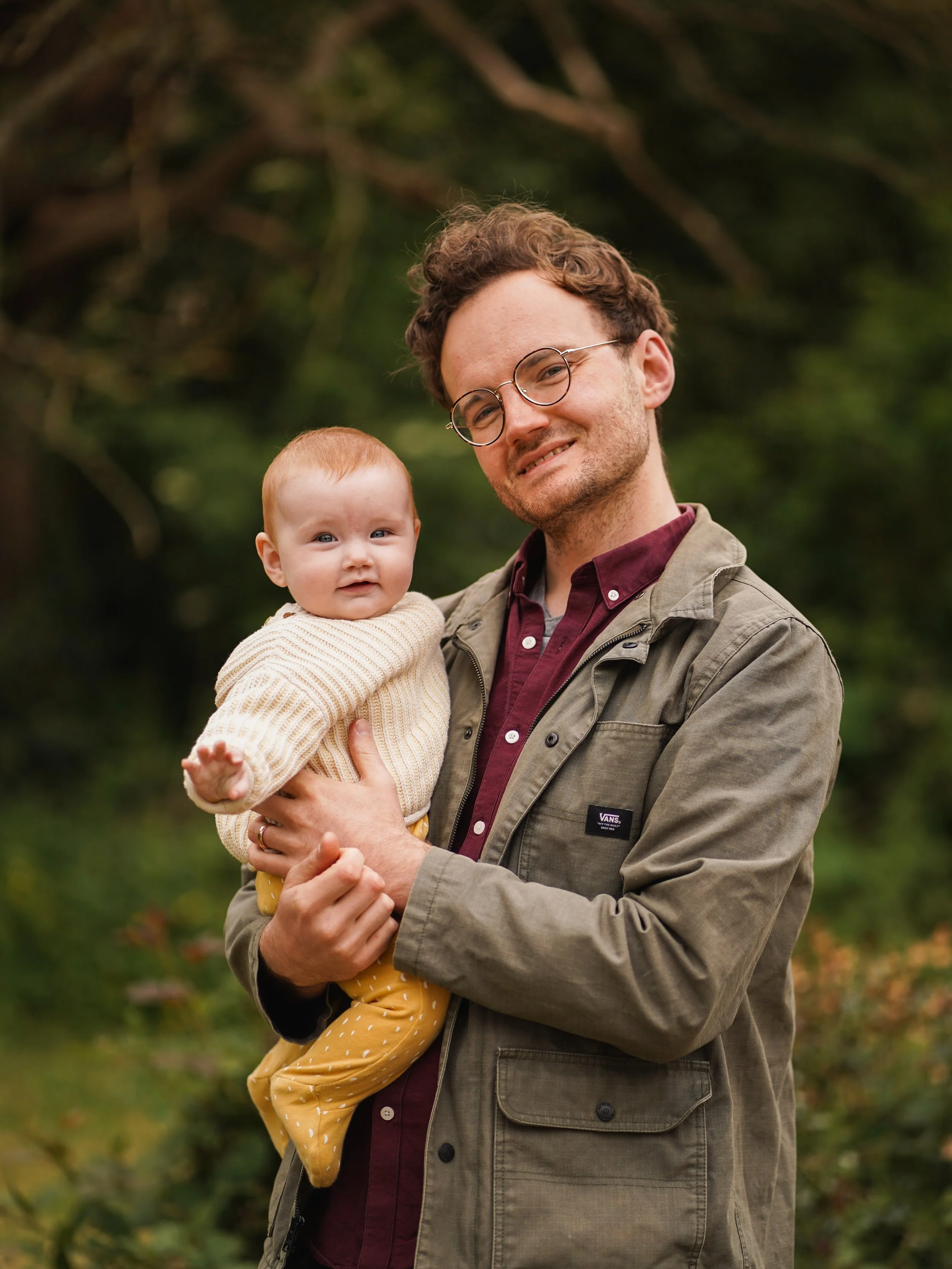 A man with curly brown hair, glasses, holding a young child with red hair and blue eyes, outdoors with blurred green foliage in the background.