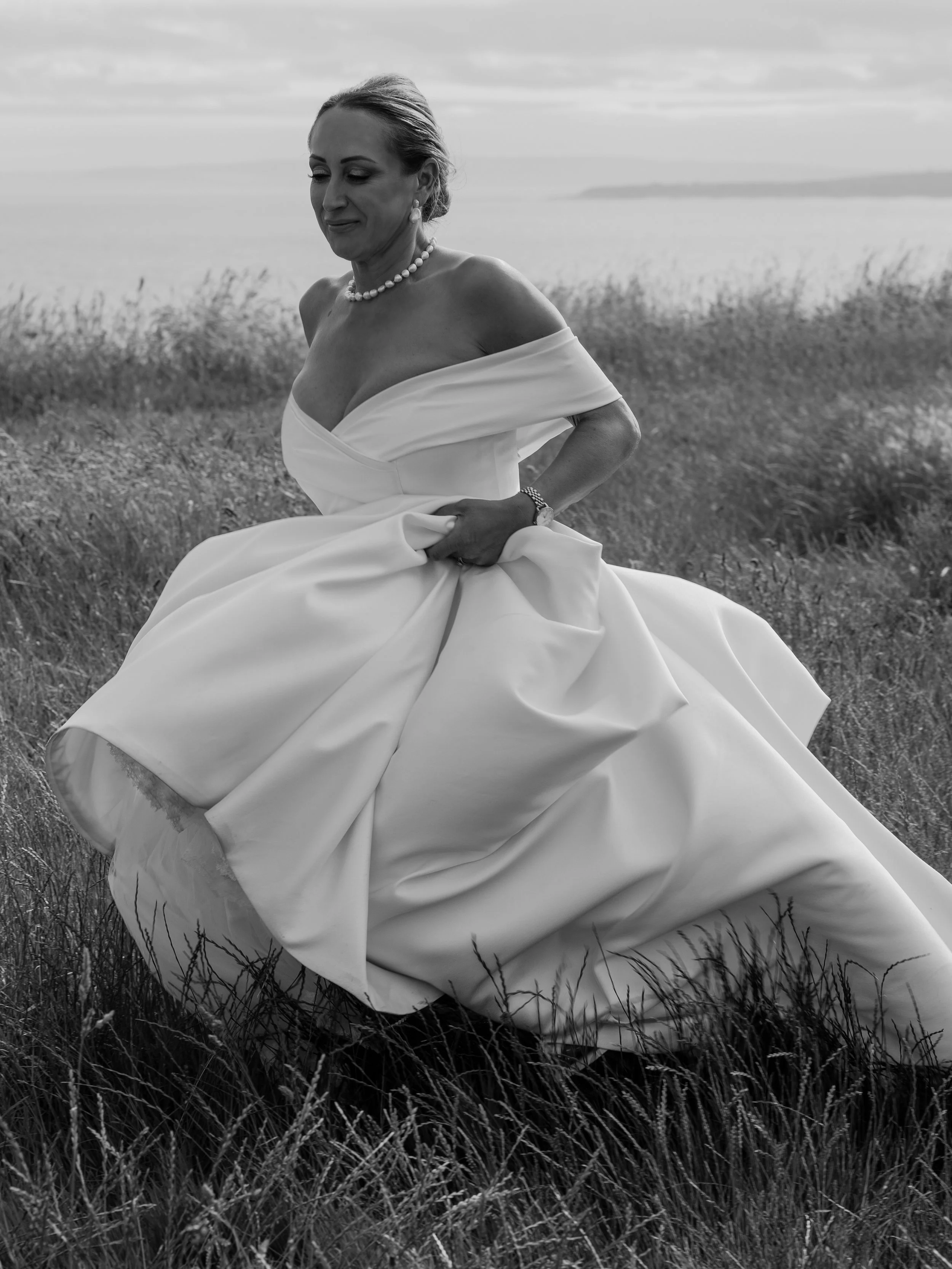 A black and white photo of an elegant woman in a wedding dress standing outdoors in a grassy field, with a body of water and cloudy sky in the background.