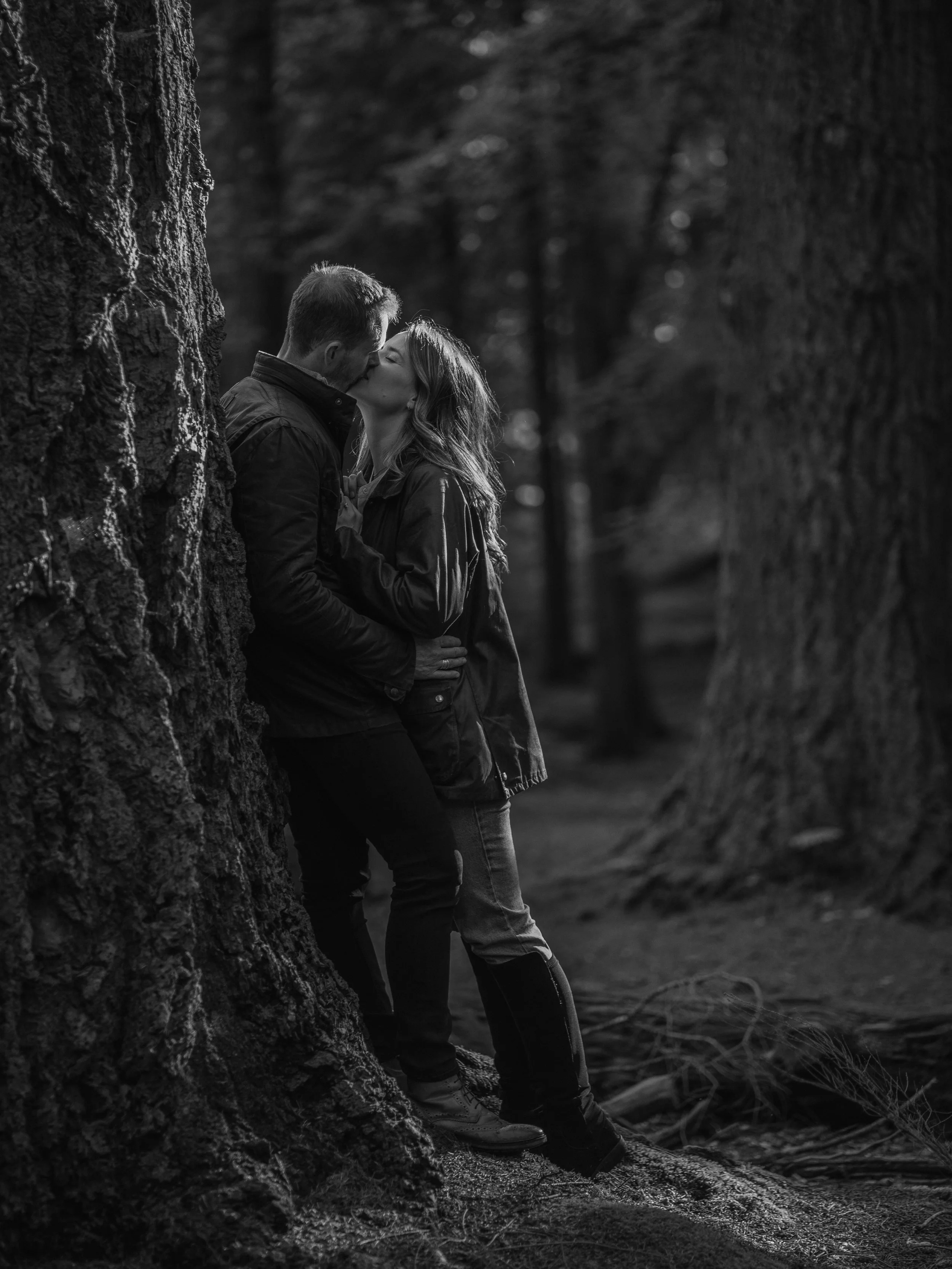 A black and white photo of a couple sharing a kiss in a forest, standing close to a large tree, with other trees and a forest path in the background.