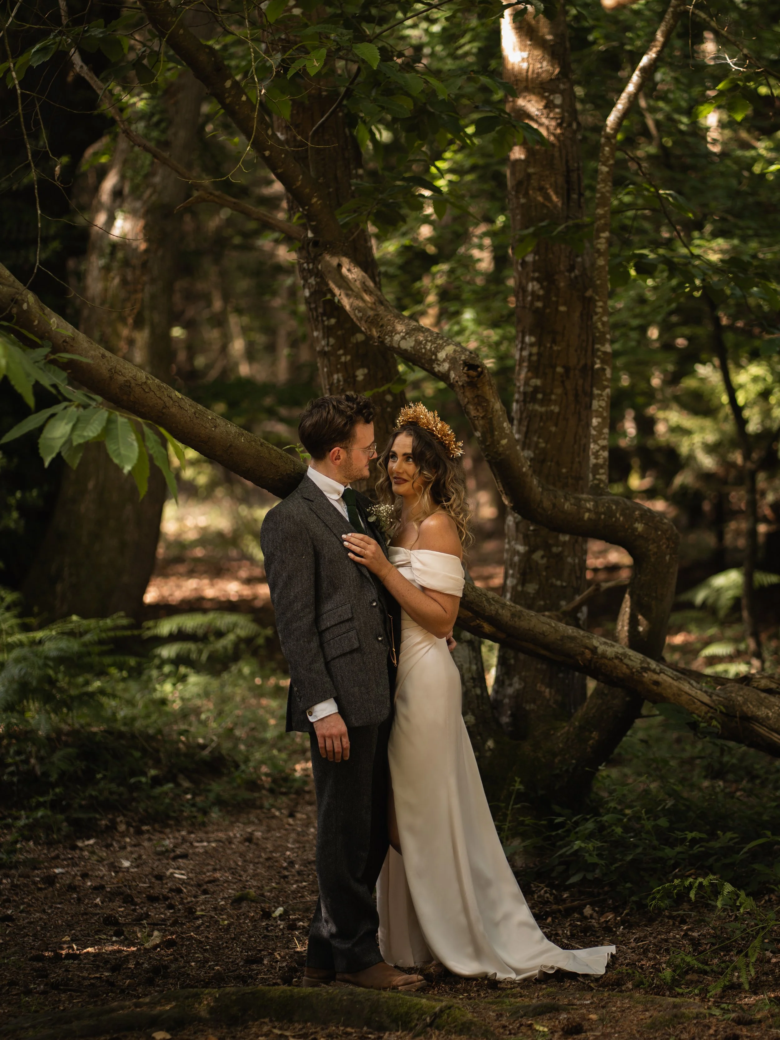 A bride and groom standing close together in a forest, facing each other. The groom wears a dark grey suit and glasses, while the bride wears an off-shoulder white wedding dress and a gold floral crown. They appear to be gazing into each other's eyes