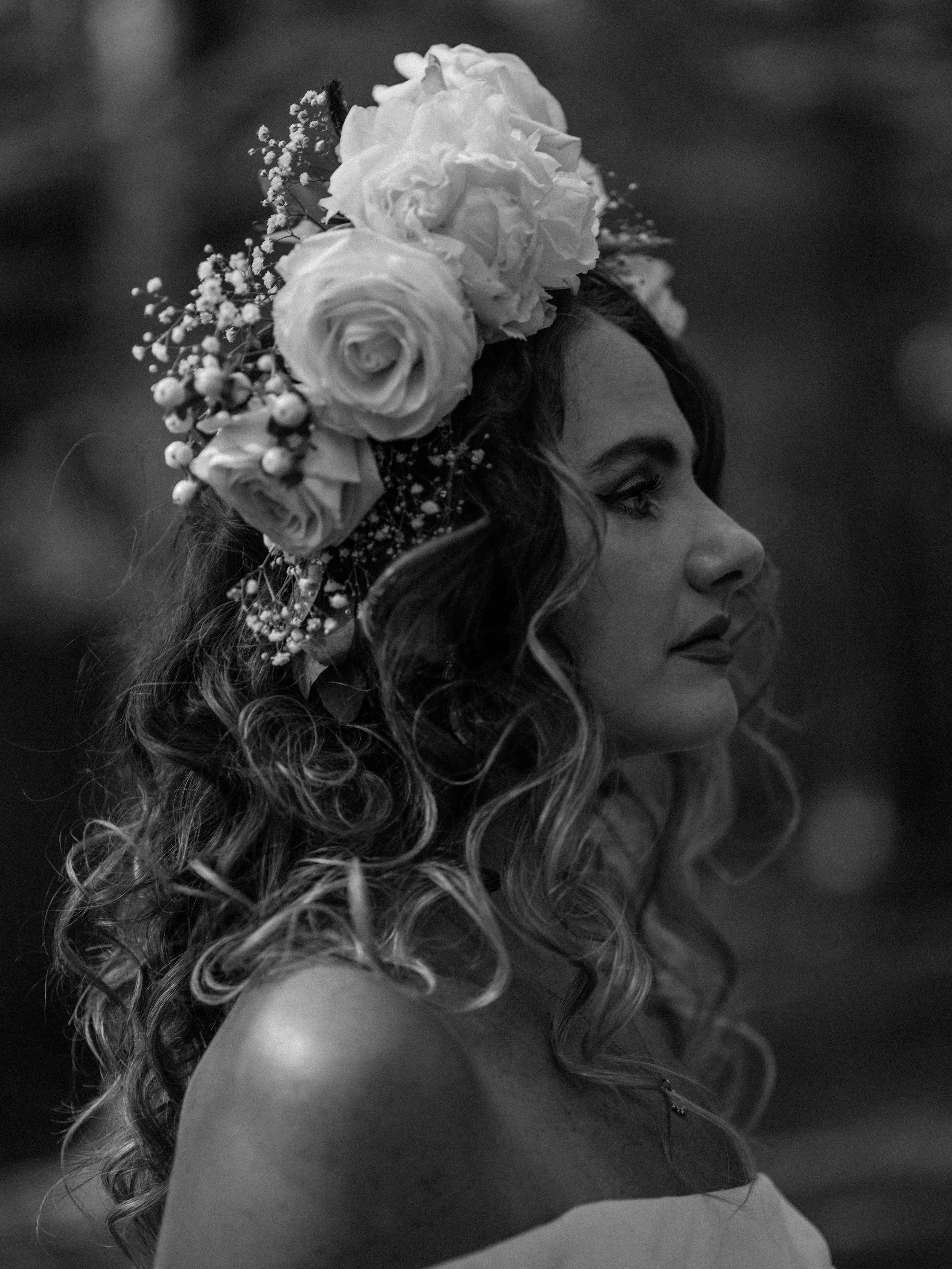 Black and white photo of a woman with curly hair wearing a headpiece made of roses and small flowers, looking to the side.