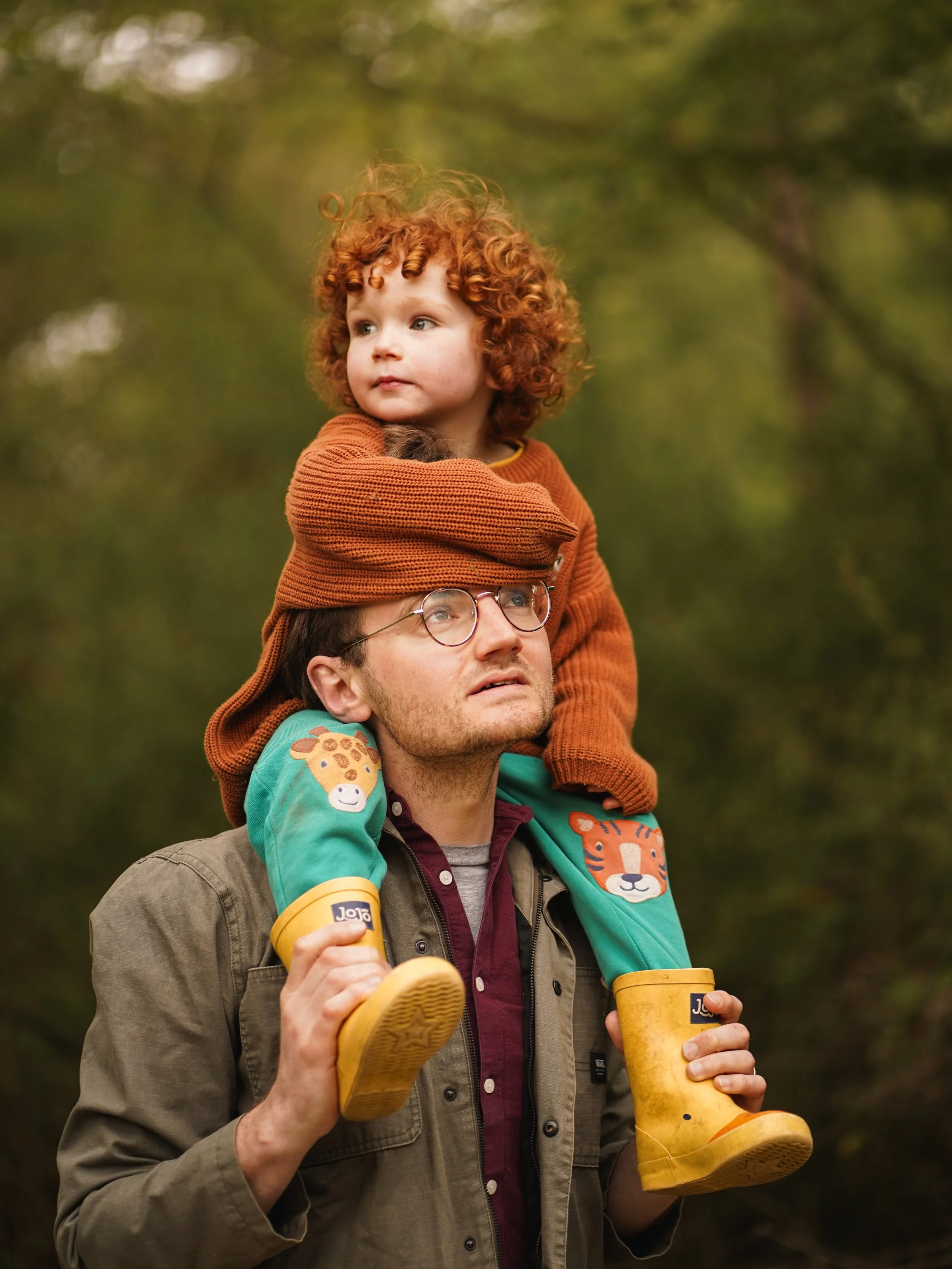 A man is carrying a young child on his shoulders. The child has curly red hair and is dressed in an orange sweater. The man is holding two yellow boots and a yellow cup, and wears glasses. The background is an outdoor scene with trees and greenery.