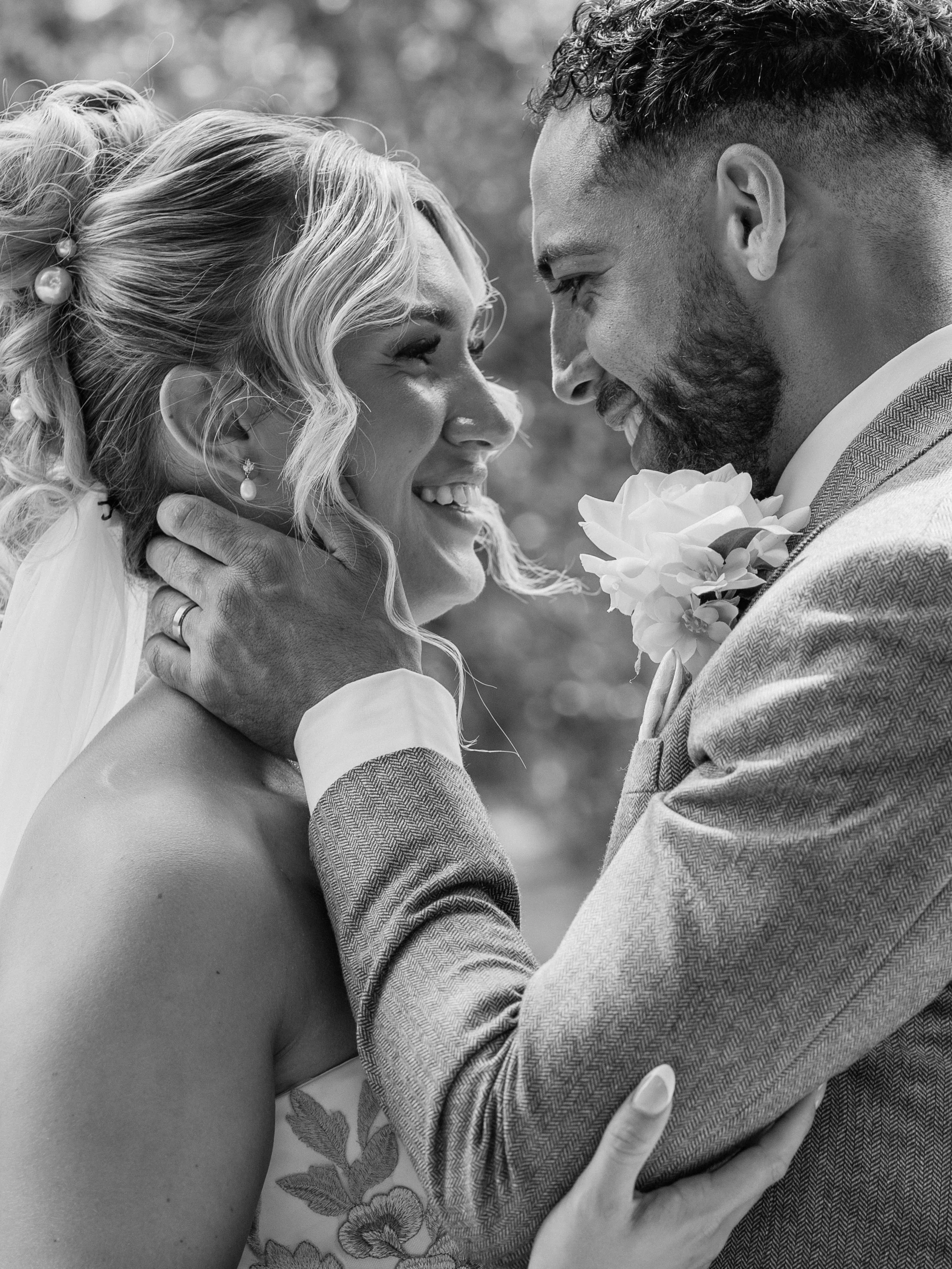 A black and white photo of a couple in wedding attire, smiling and touching foreheads closely, with a blurred outdoor background.