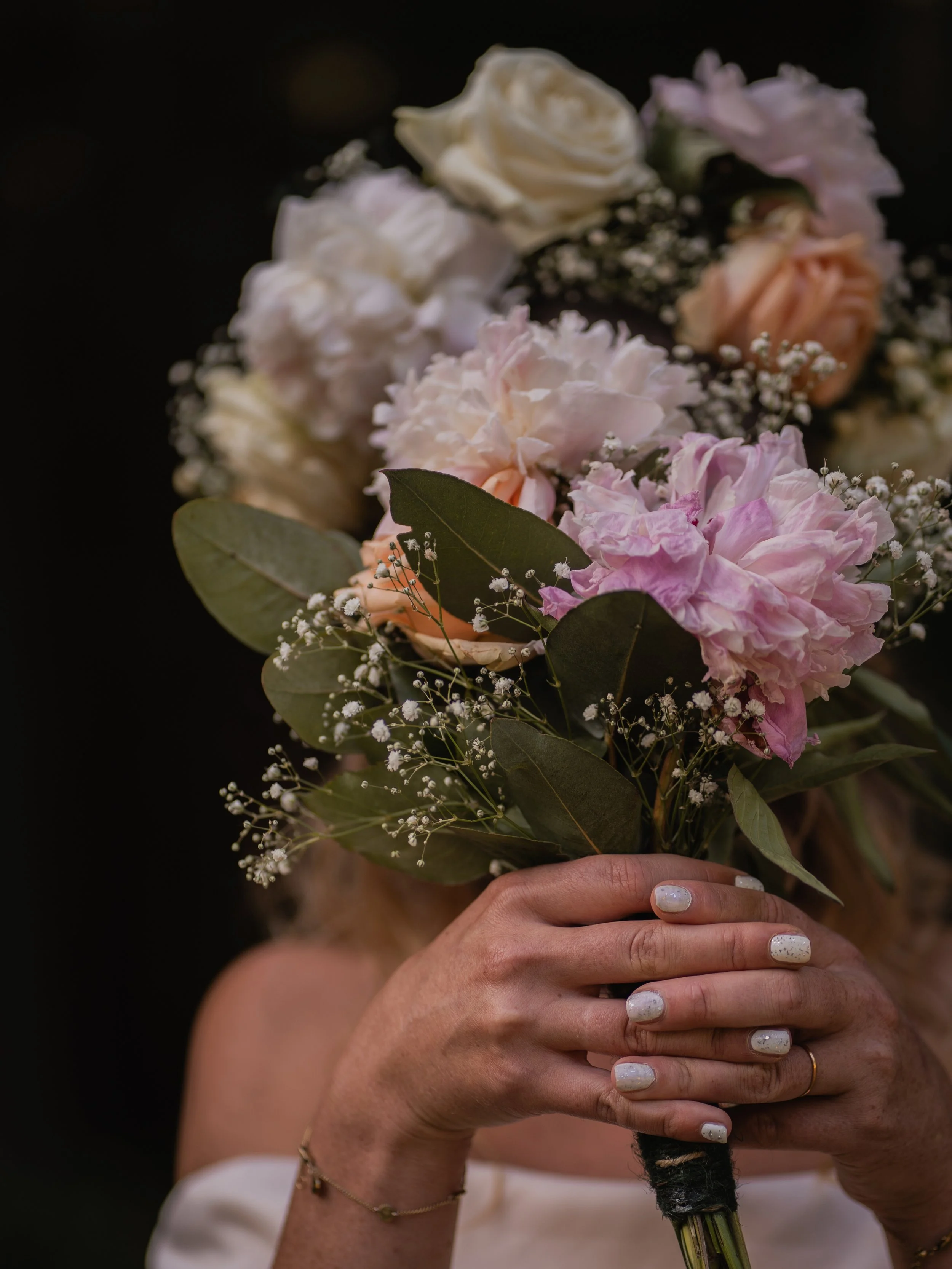 Close-up of a person holding a bouquet of mixed pastel-colored roses and flowers, with a dark background, showing hands and part of the person's arm with painted nails and jewelry.