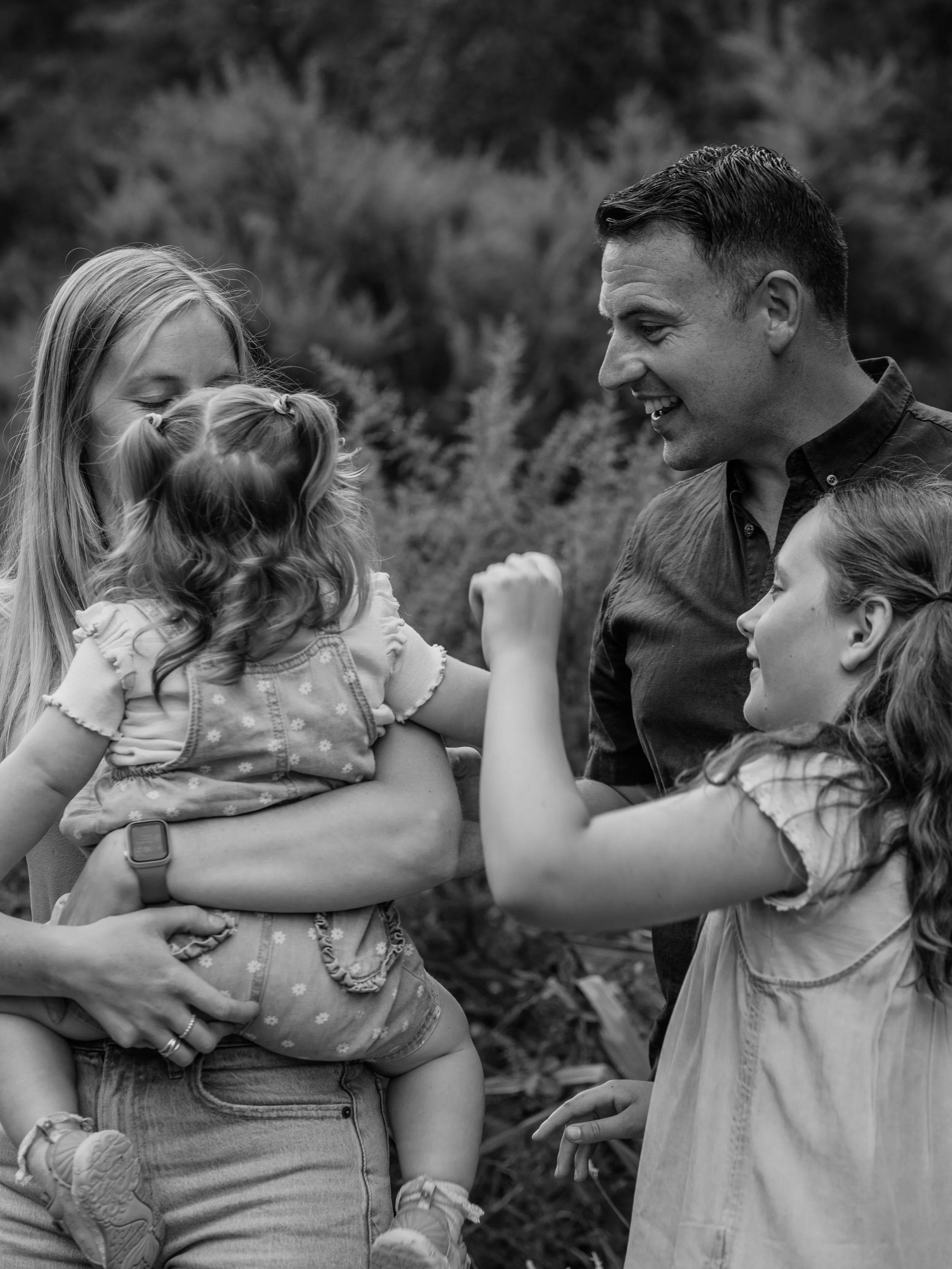 A black and white photograph of a family outdoors. The woman is holding a young girl, and both are smiling. A man and another girl are facing them, smiling and engaging in a playful moment.