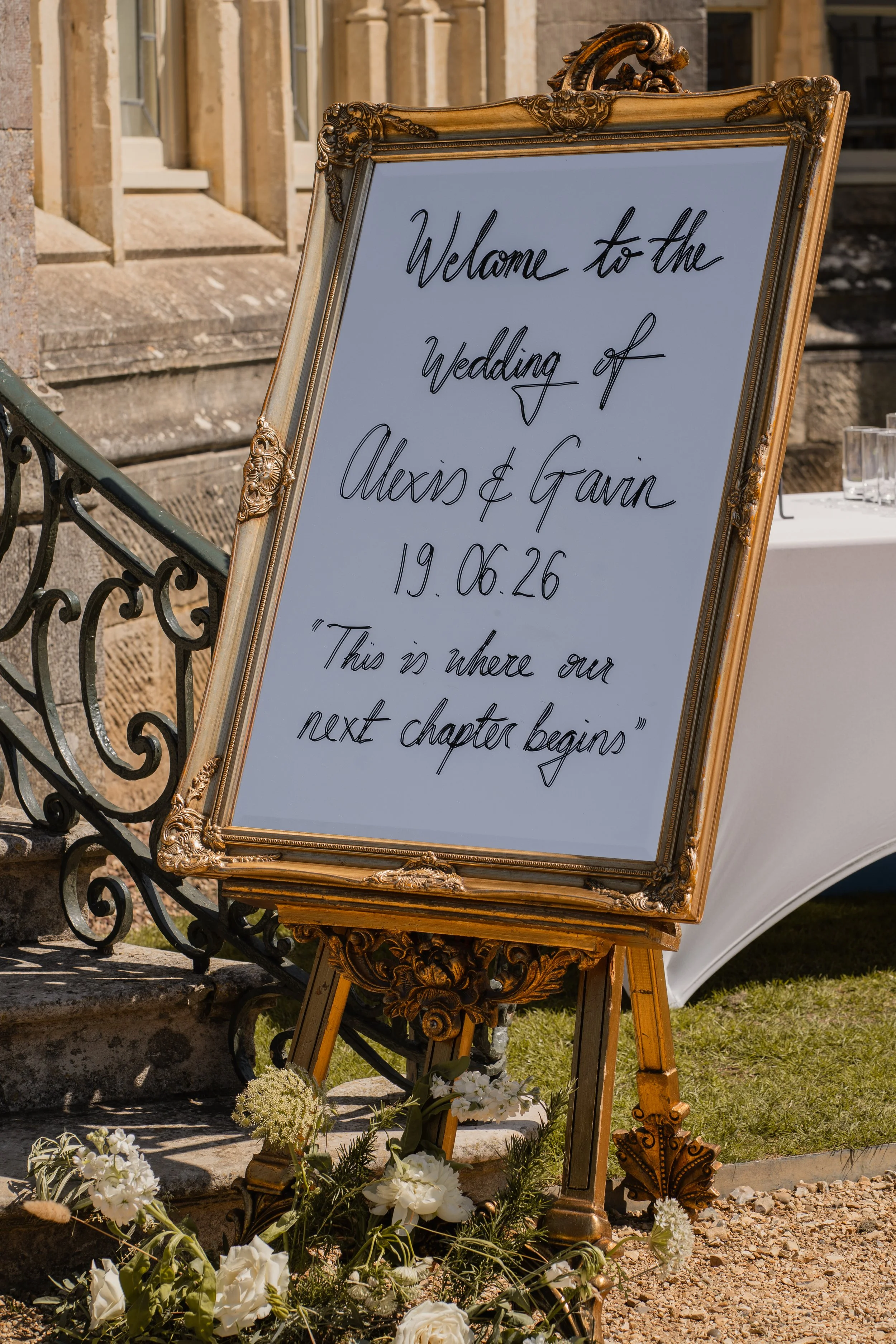 A decorative wedding welcome sign with ornate gold frame, displayed outdoors on an easel. The sign reads, 'Welcome to the Wedding of Alex & Gavin 19.06.26. Below the sign are white flowers and greenery. The background includes stone steps, a metal ra