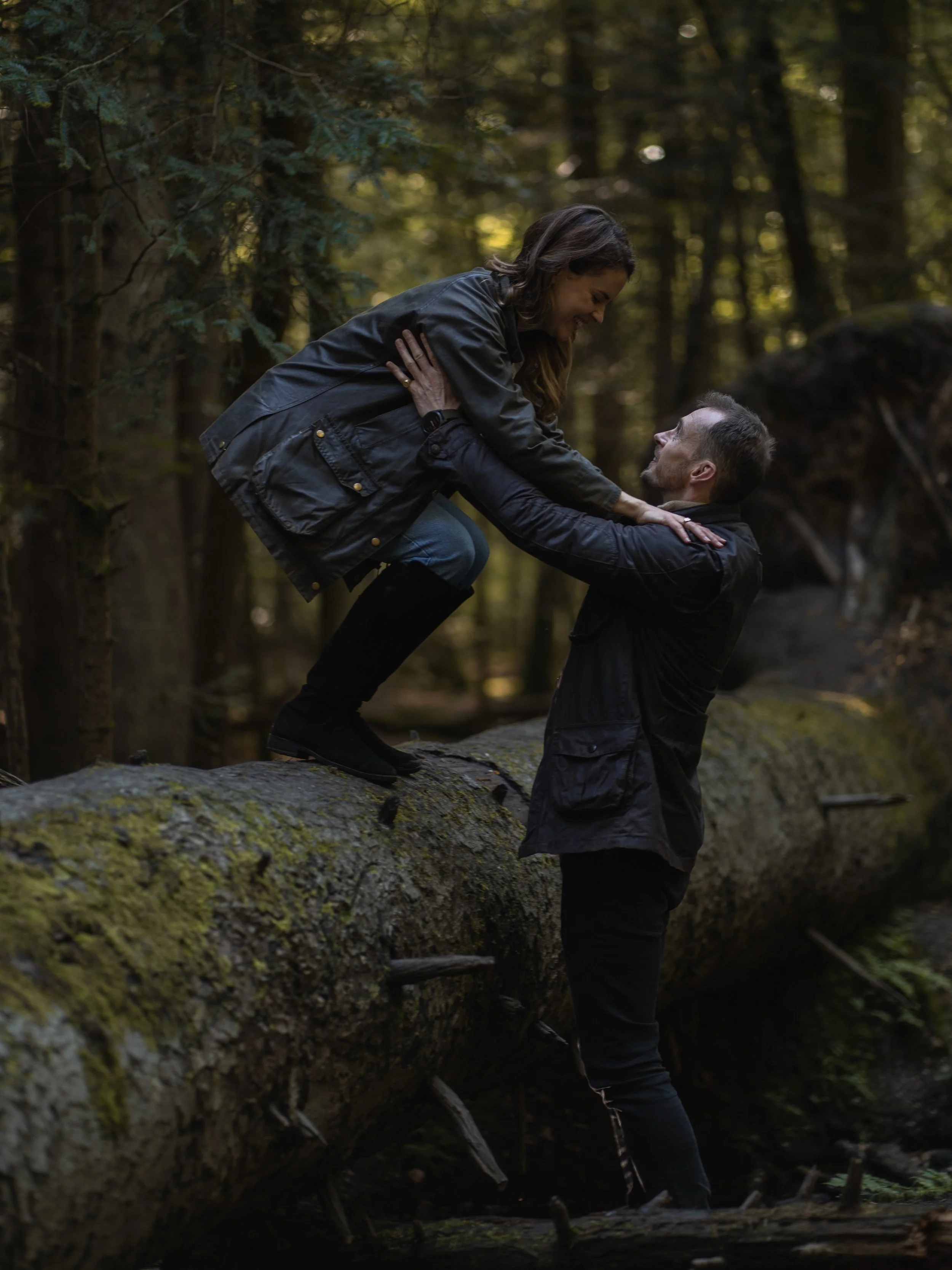 A man and woman sparring outdoors in a forest, with the woman being lifted by the man over fallen tree trunks.