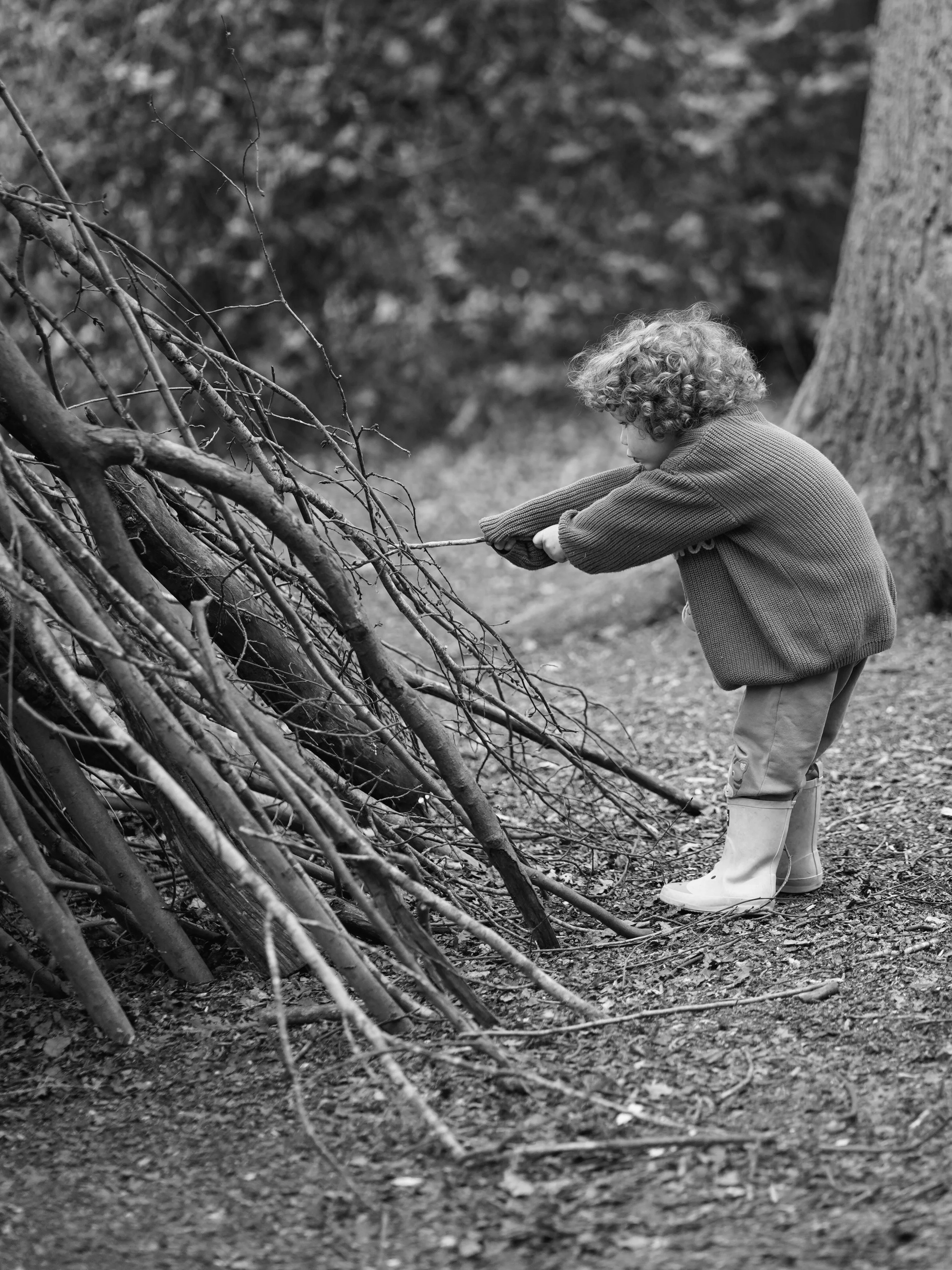 A young child with curly hair wearing a sweater, pants, and boots, bends over outdoors near a large tree, picking up a stick from a pile of sticks and branches.