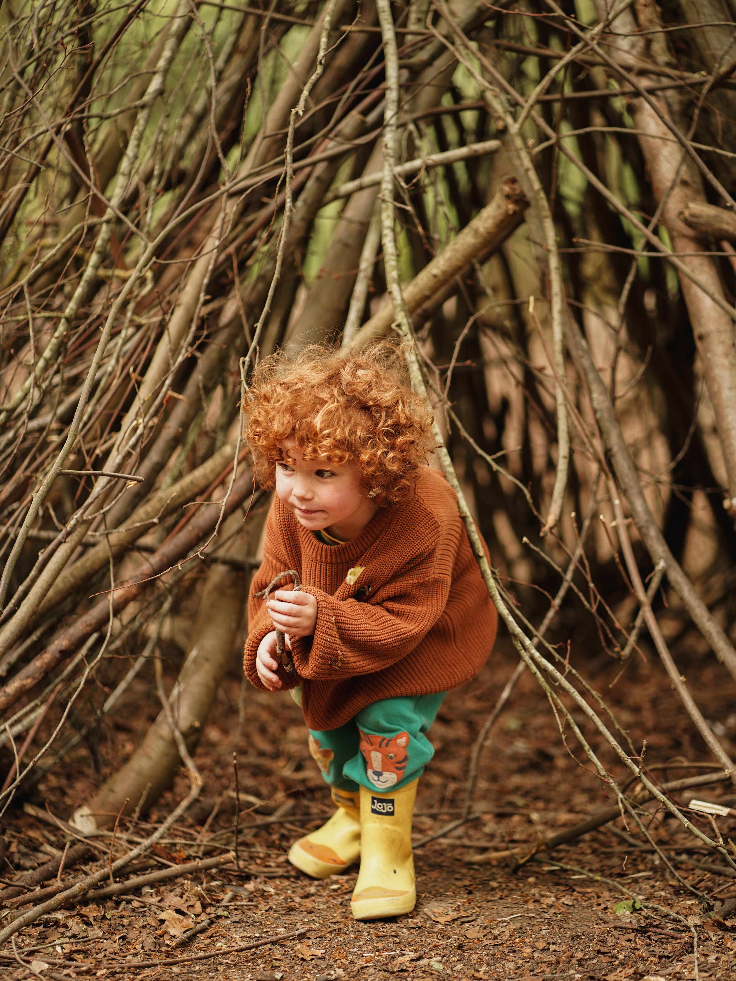 A young child with curly red hair wearing a brown sweater, green pants with a lion face on the knee, yellow rain boots, and yellow rain boots, crouching inside a small shelter made of sticks and branches in a wooded area.