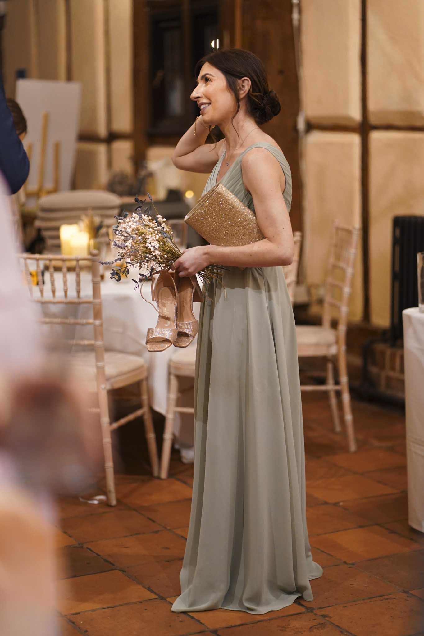 A woman in an elegant, sleeveless, light green gown holding a bouquet of flowers and a sparkly gold clutch, at what appears to be a formal event or wedding reception, standing on a wooden floor with chairs and candles in the background.